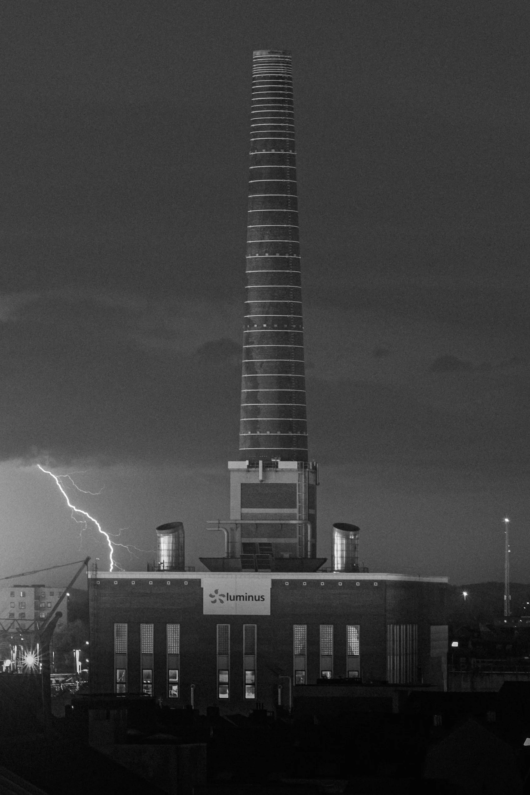 Nighttime photo of a tall industrial smokestack with lightning in the background and a sign that says 'Luminus' on a building at the bottom.