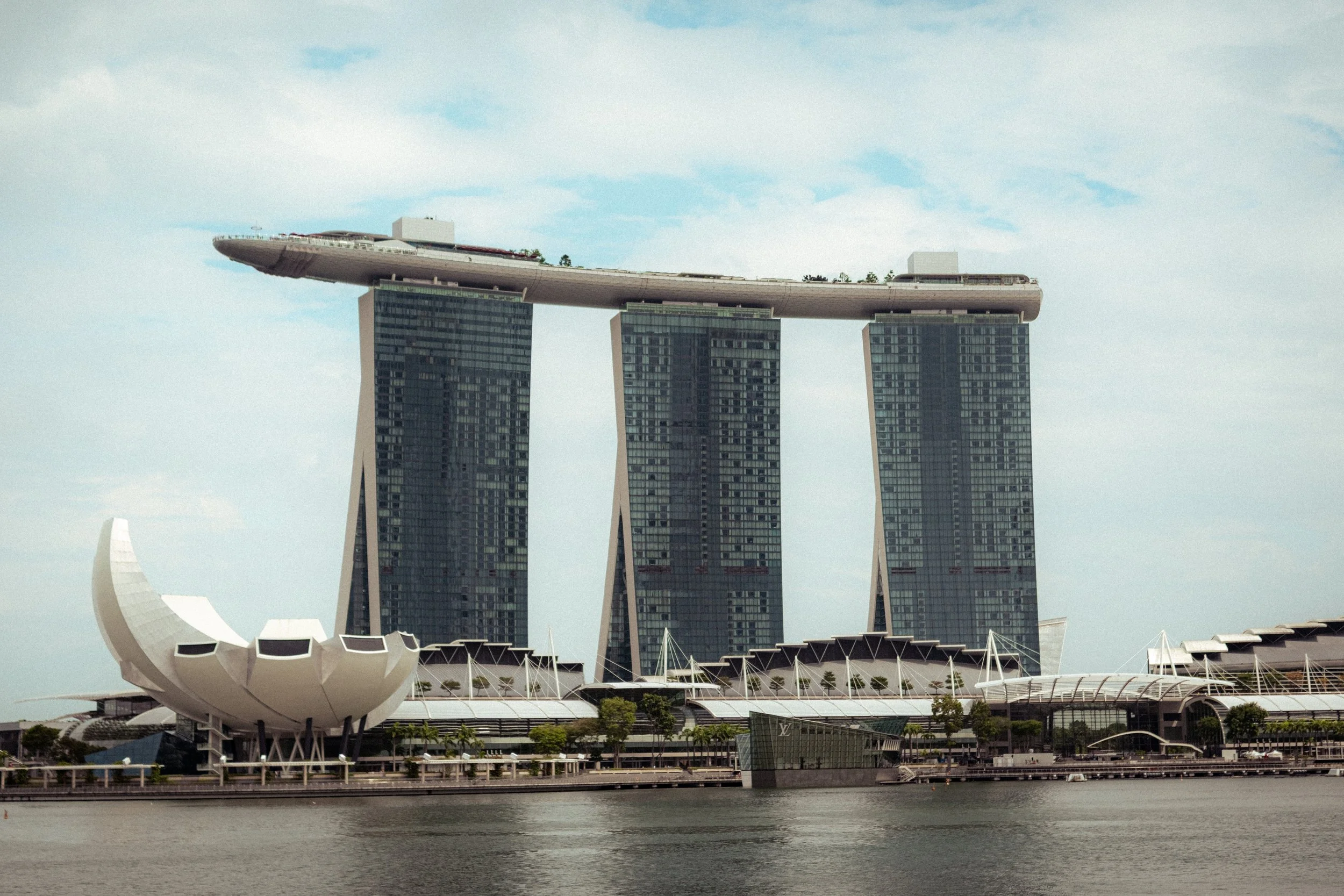Singapore's Marina Bay Sands hotel with three tall towers and a boat-shaped structure on top, along with the ArtScience Museum with its lotus petal design, viewed from across the water.