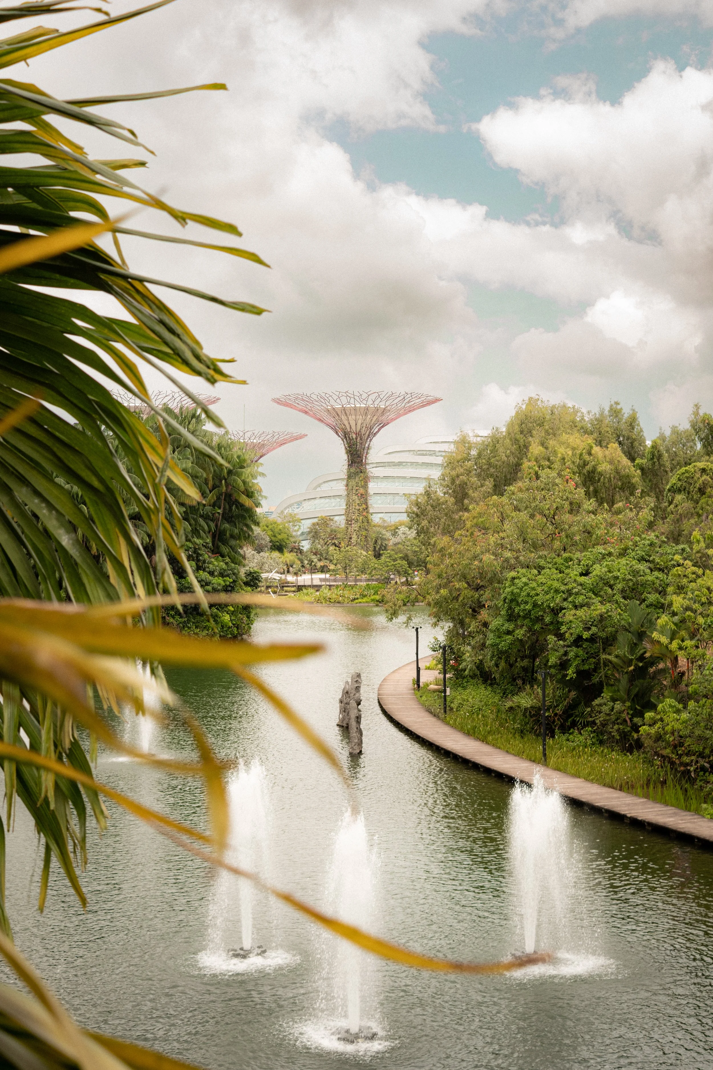 A scenic park with a narrow lake, a curved walkway on the right, lush trees on the right, and water fountains shooting water into the lake. In the background, large futuristic structures and two tall, distinctive Supertree Grove structures are visibl