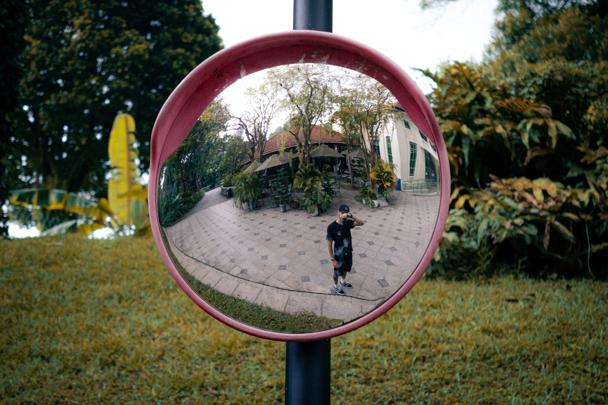 Reflected image of a person taking a photo in a circular mirror outside, with trees and a building in the background.