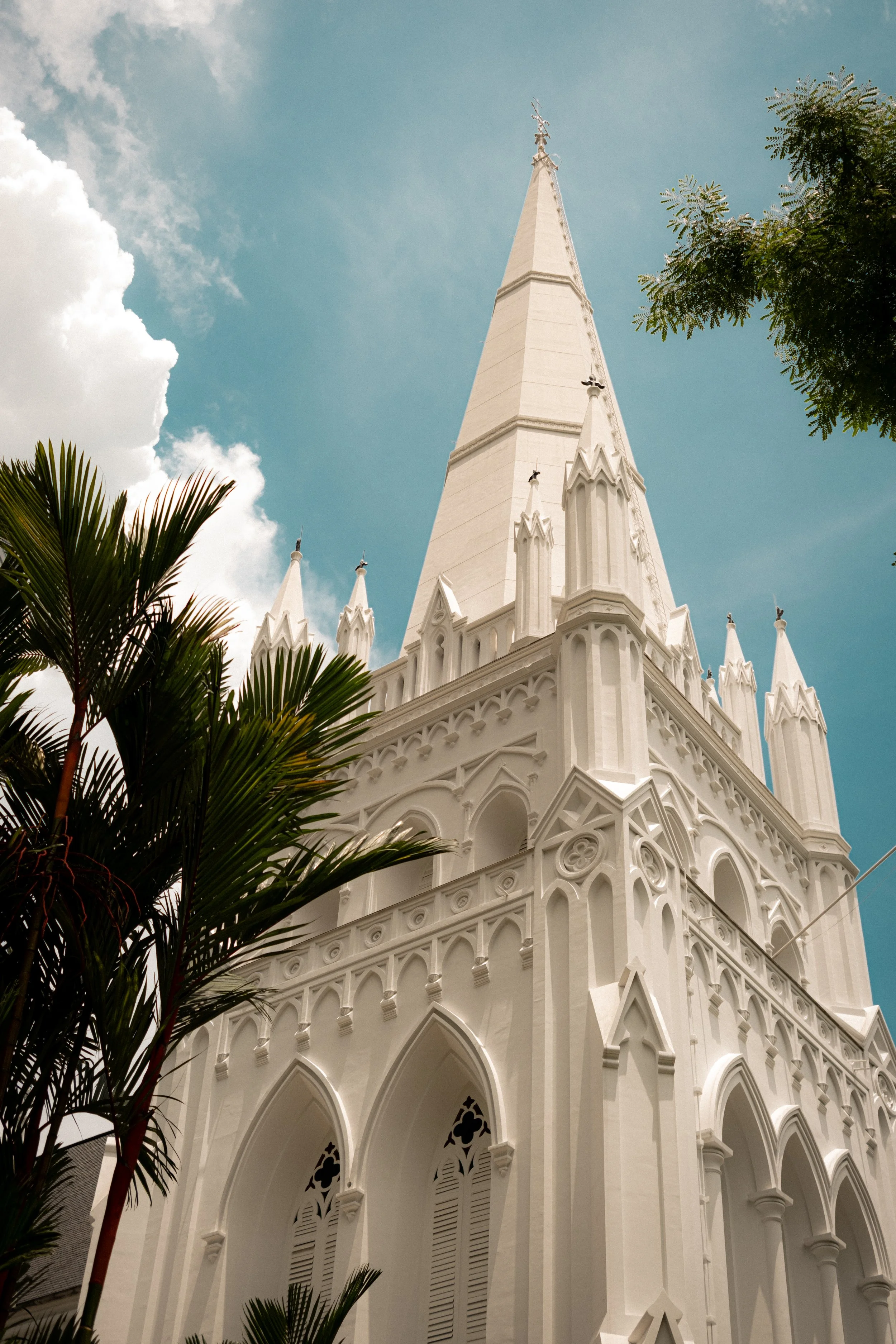 A white Gothic-style church with pointed arches, intricate details, and a tall steeple, seen from below, with green palm trees in the foreground and a partly cloudy sky in the background.