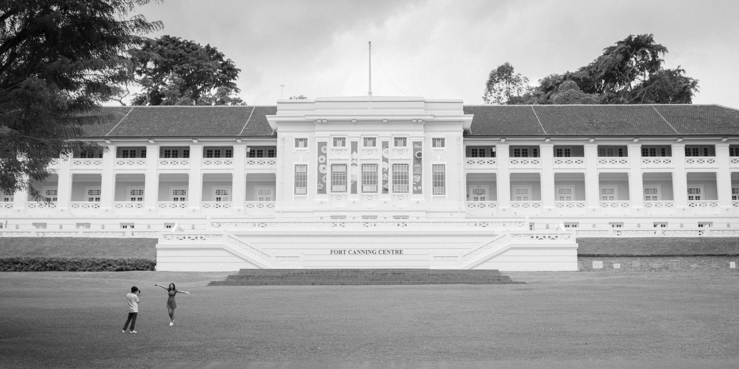 Black and white photo of a historic white building with columns labeled 'Fort Canning Centre' in Singapore, with children and adults playing on a lawn in front.