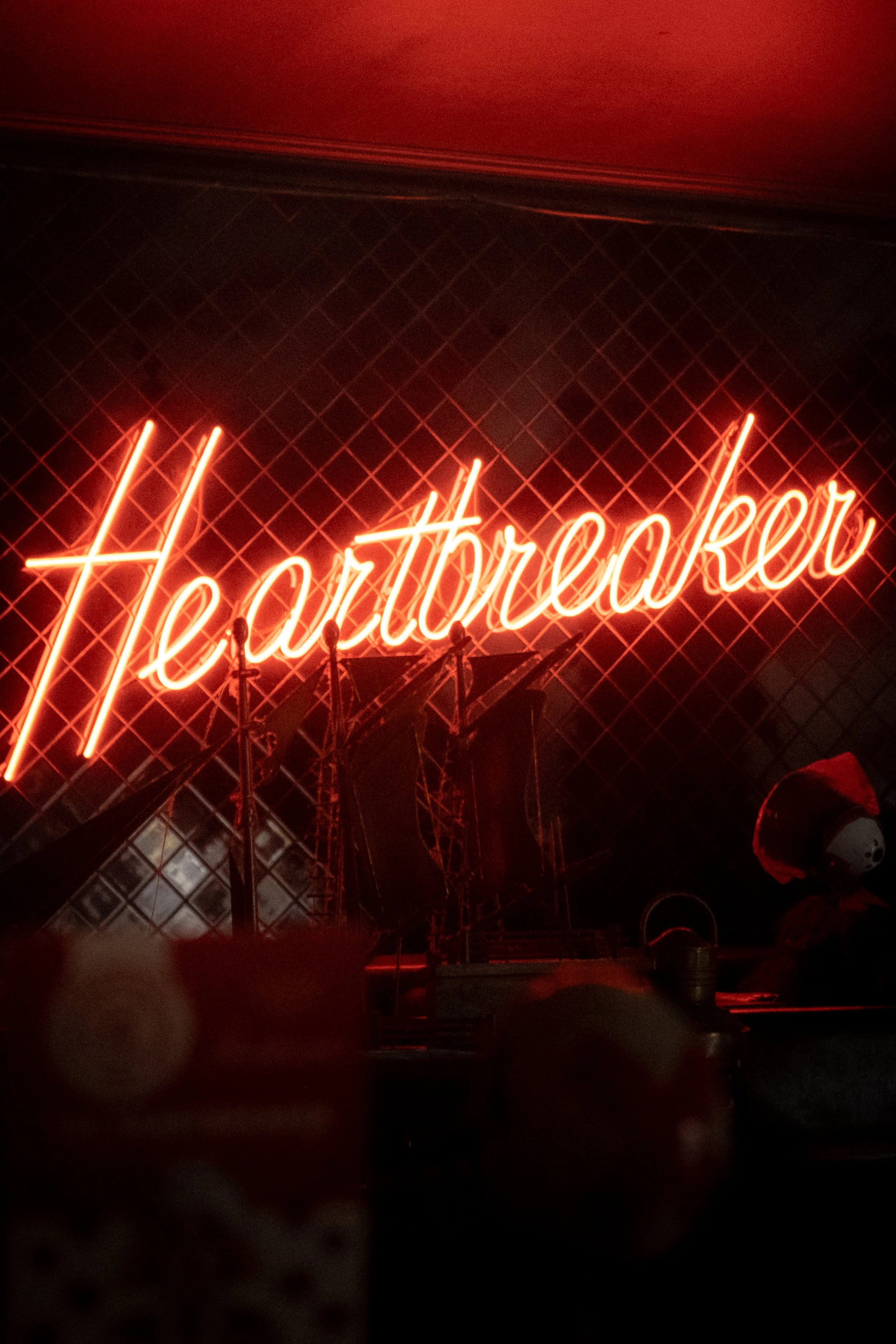 A neon sign spelling 'Heartbreakers' in cursive red on a black grid background, with stage equipment and a partially visible person in the foreground.