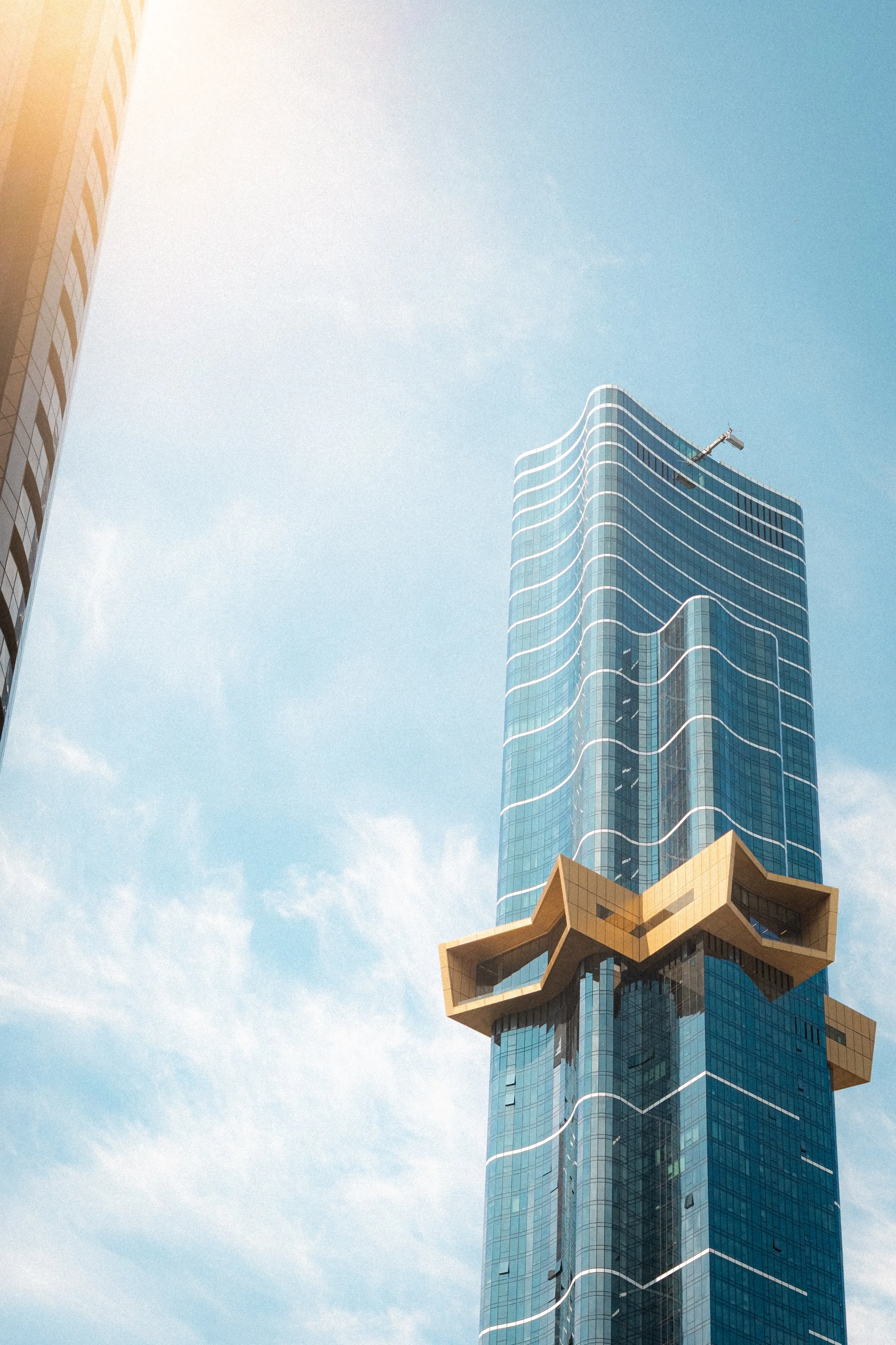 A modern skyscraper with a unique architectural design, featuring a blue glass facade with white accents and an irregularly shaped beige overhang near the top, under a light blue sky.