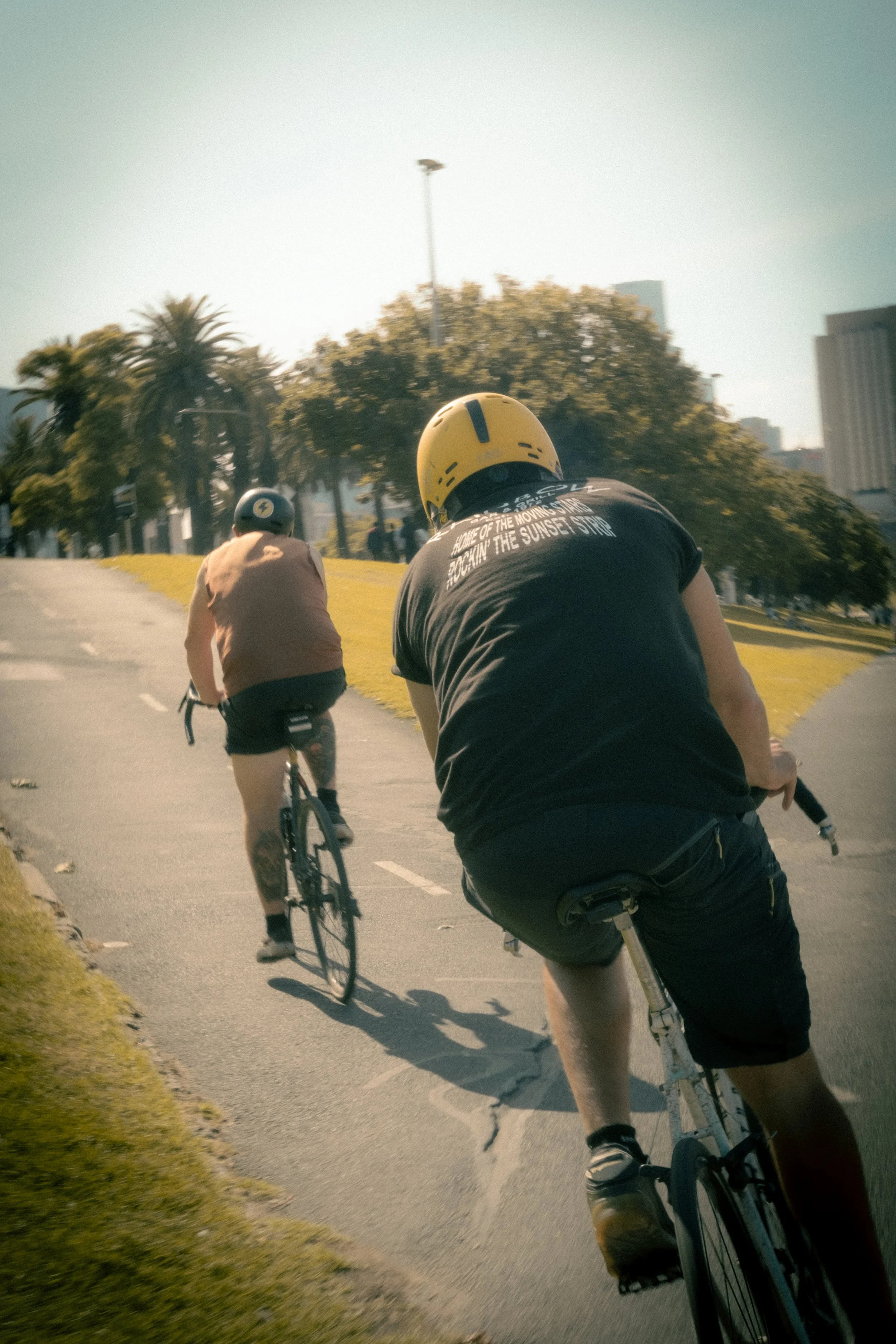 Two cyclists riding on a paved path in an urban park area with trees and tall buildings in the background under a clear sky.