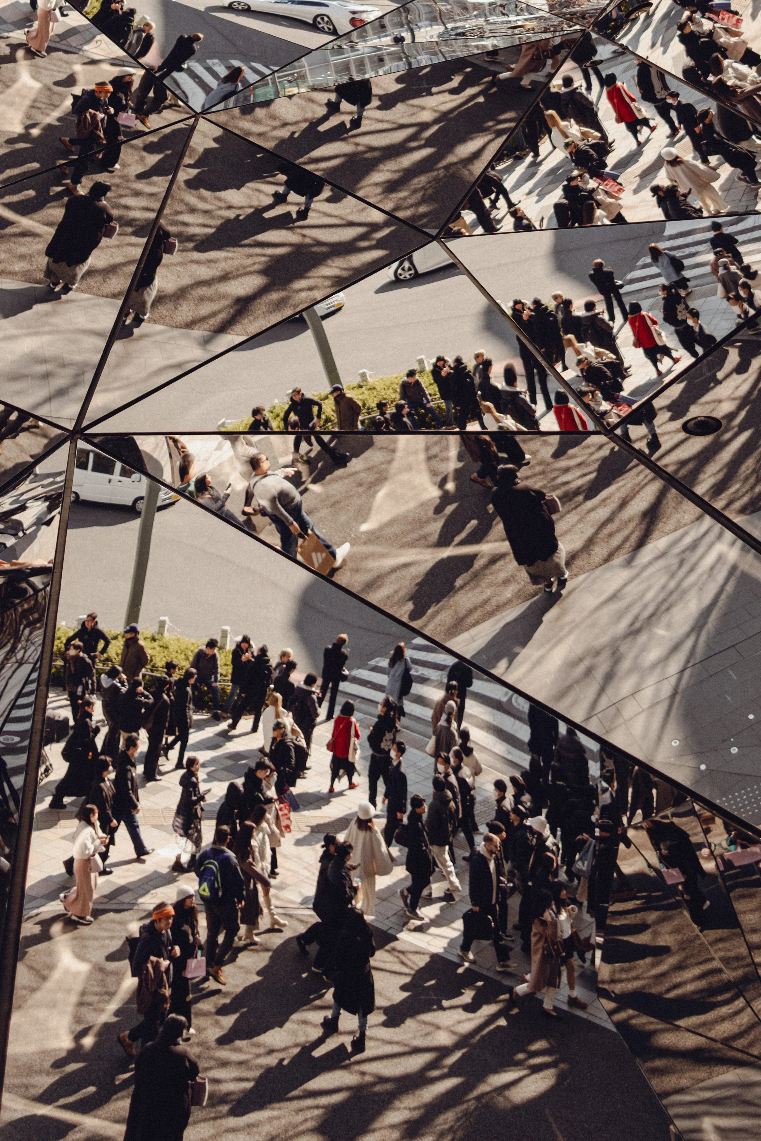 Multiple reflections of a busy city street with pedestrians walking on crosswalks and sidewalks, captured in geometric mirrored panels that create a fragmented view of the scene.