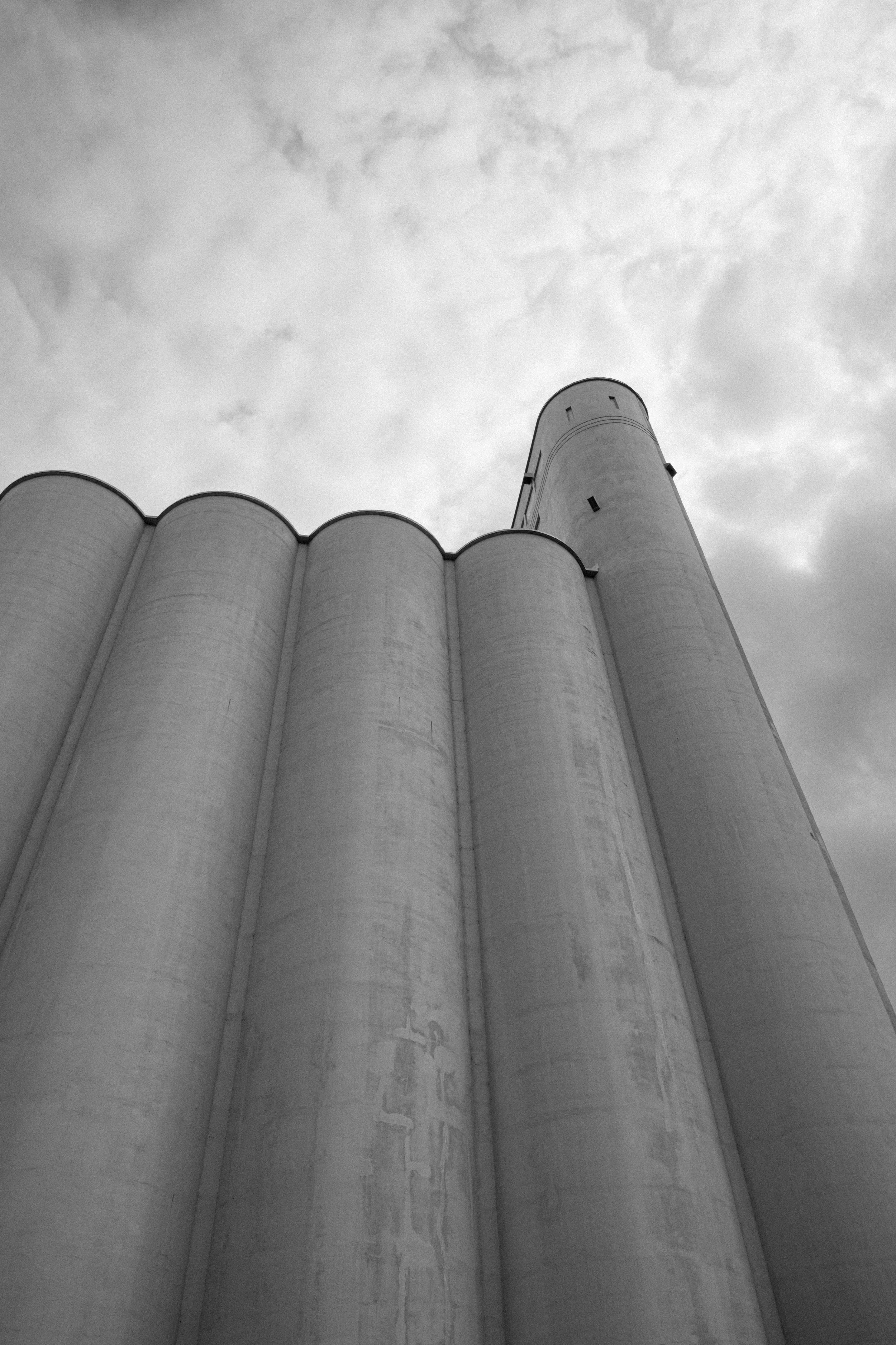 Black and white photograph of a large industrial silo or grain storage facility taken from a low angle, with a cloudy sky in the background.