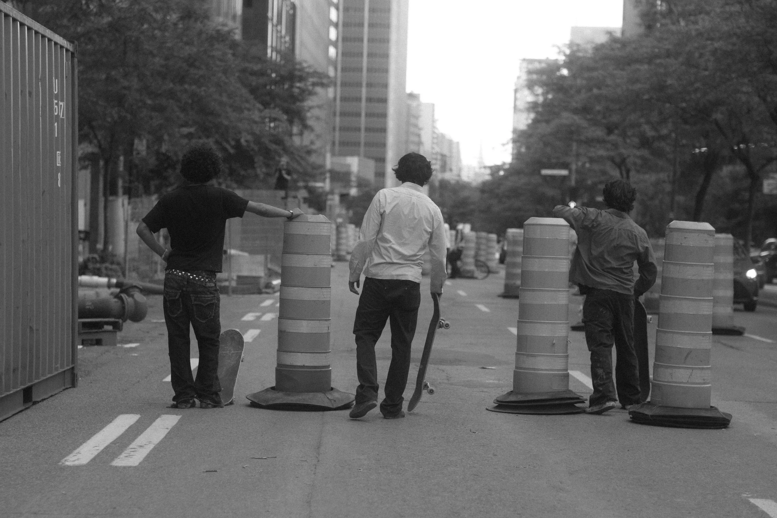 Three young men are standing on a city street, with two of them resting their arms on large traffic barrels. The street is lined with trees and tall buildings, and the photo appears to be taken during the day.