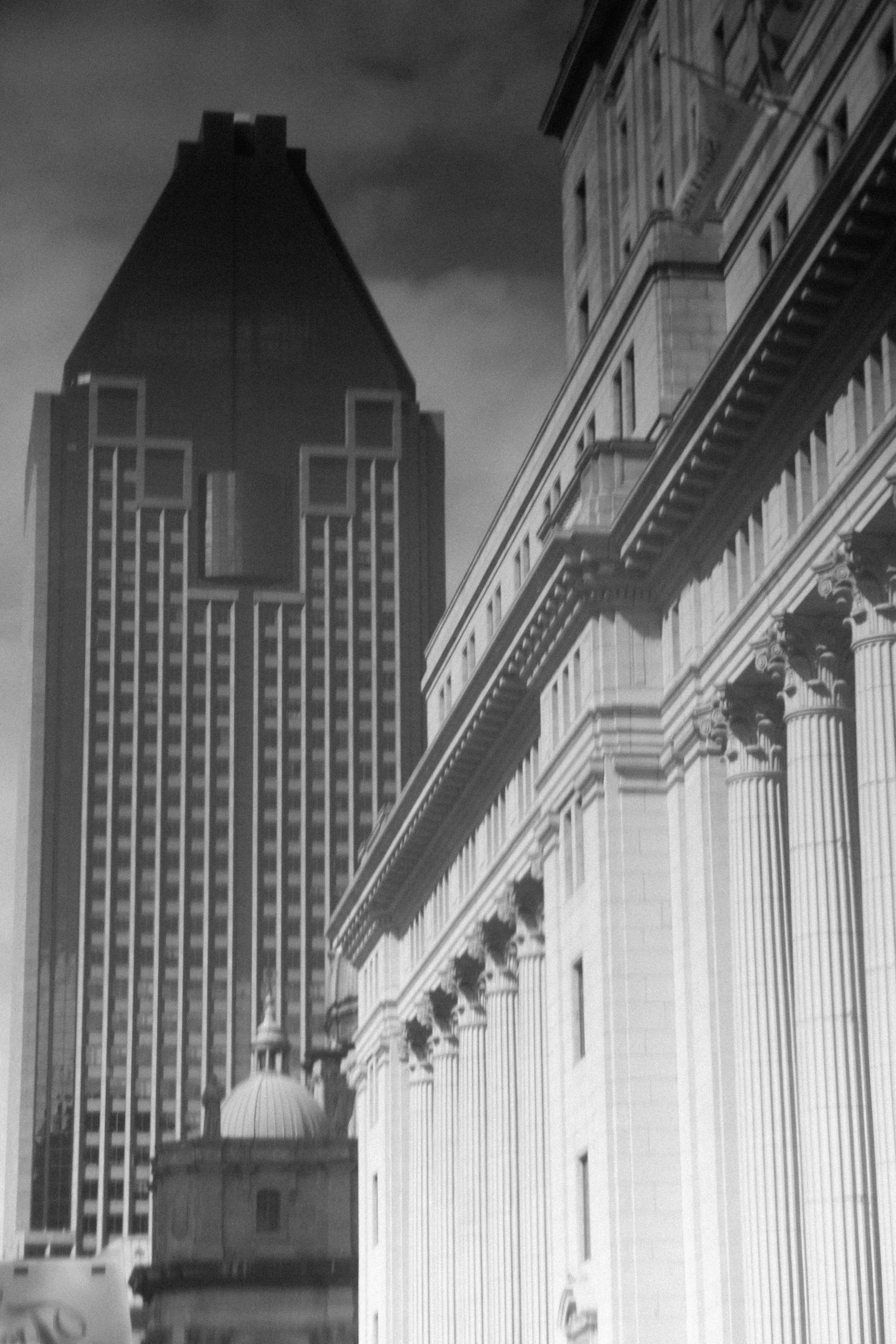 Black and white photo of city skyscrapers, one with a triangular top and multiple windows, and classical building elements on the right with columns and decorative features.