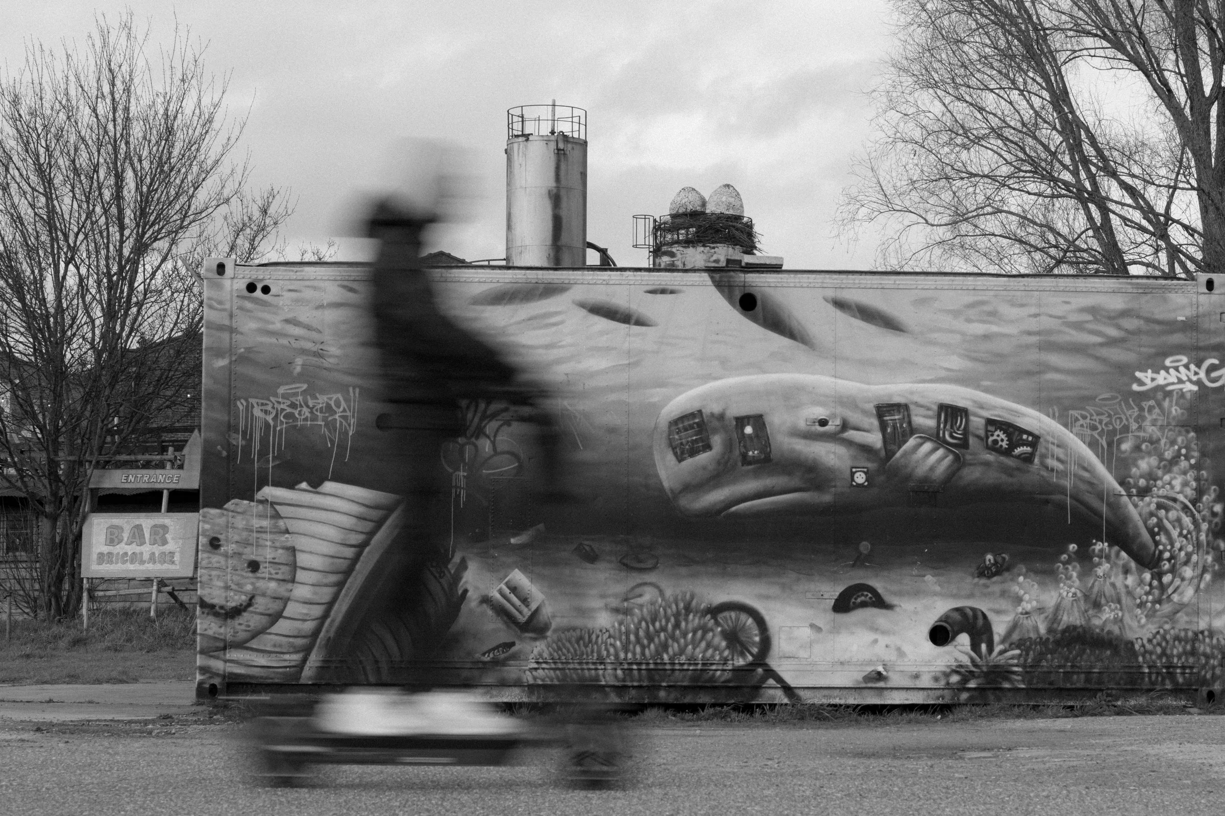 Black and white photo of a person walking past a graffiti-covered truck with a mural of a dinosaur, trees in the background, and cloudy sky.