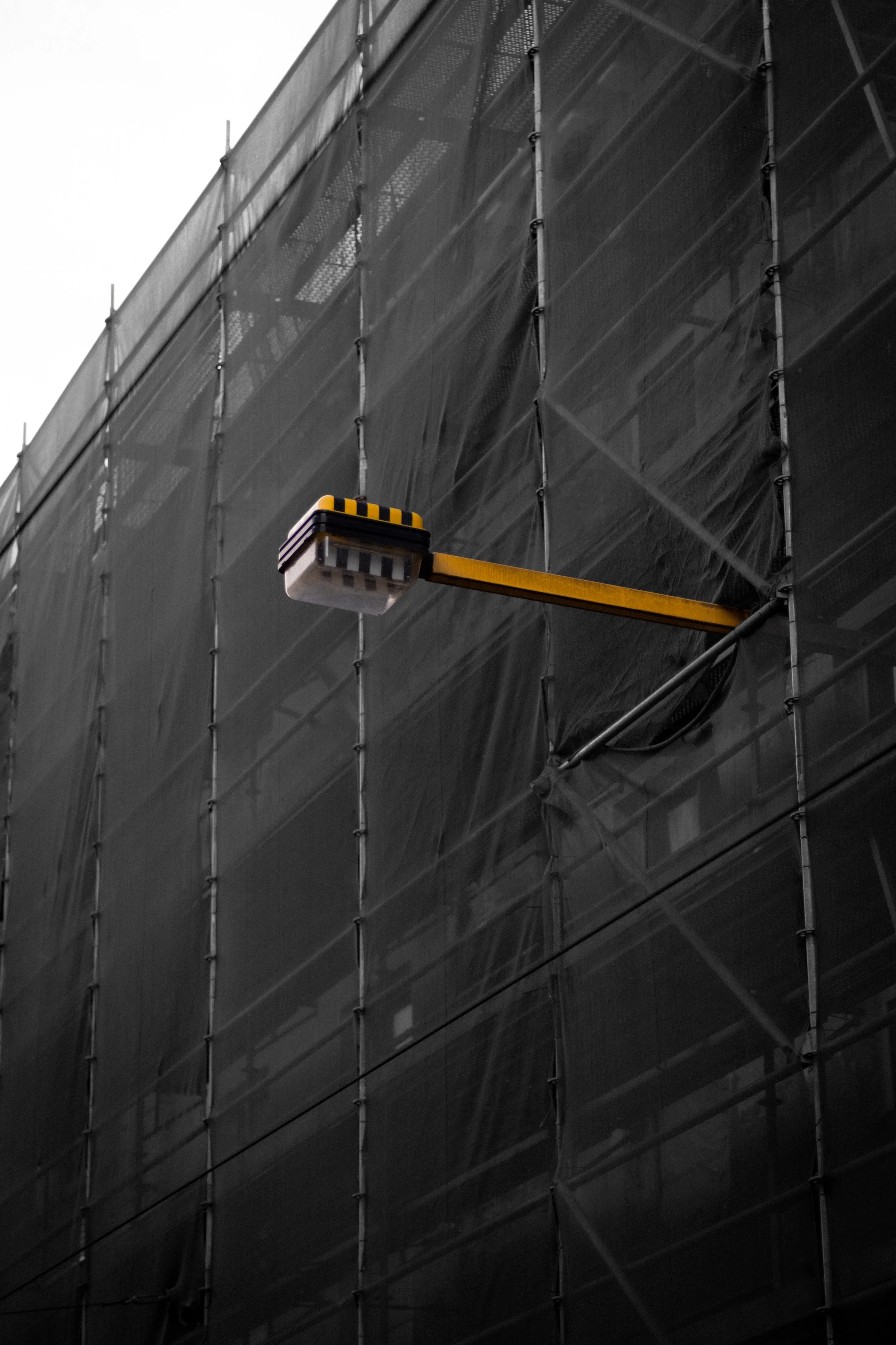 A building covered with scaffolding and black protective netting, with a suspended yellow and black safety fixture.