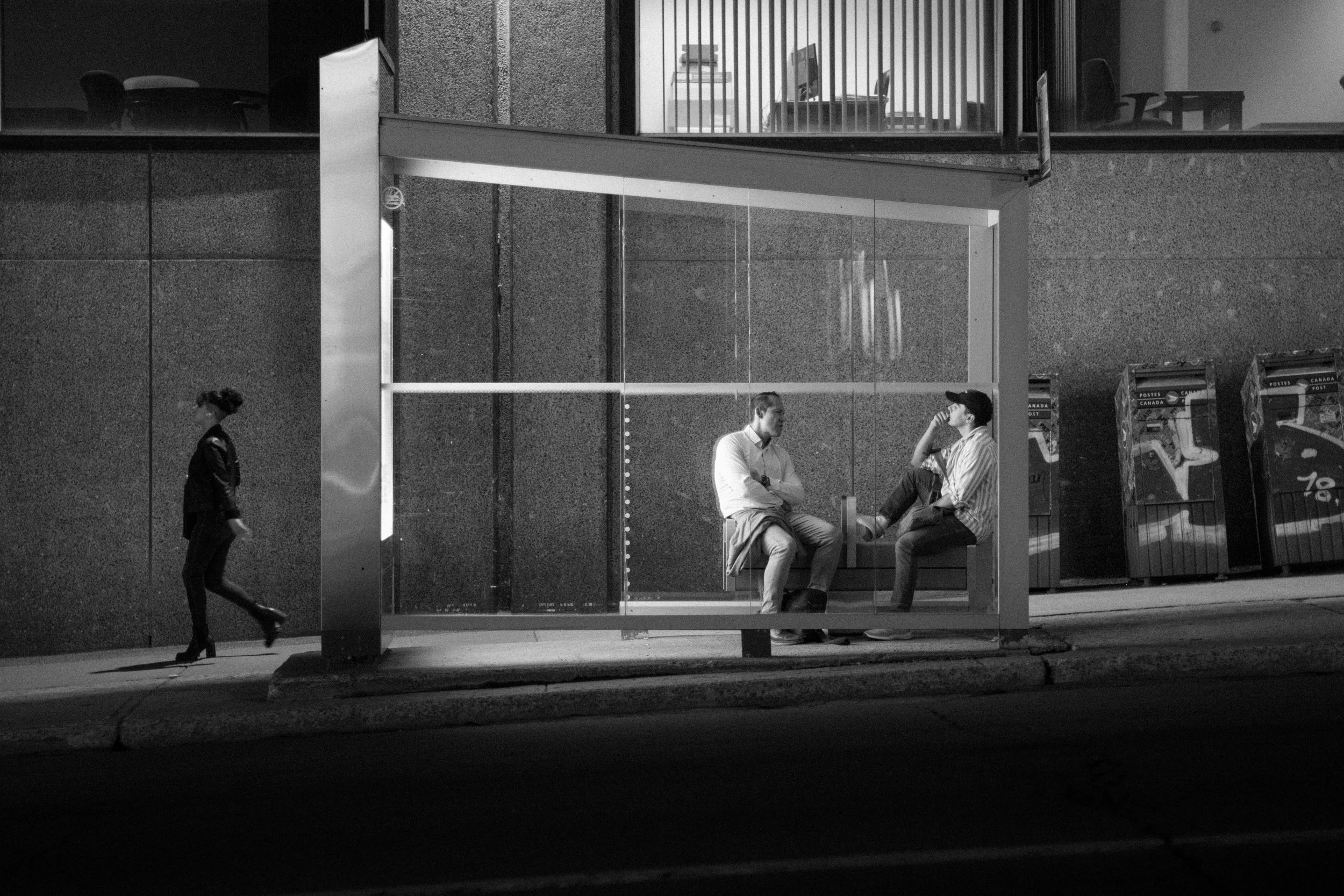 A black and white photo of a city bus stop with two men sitting on a bench and a woman walking past nearby.