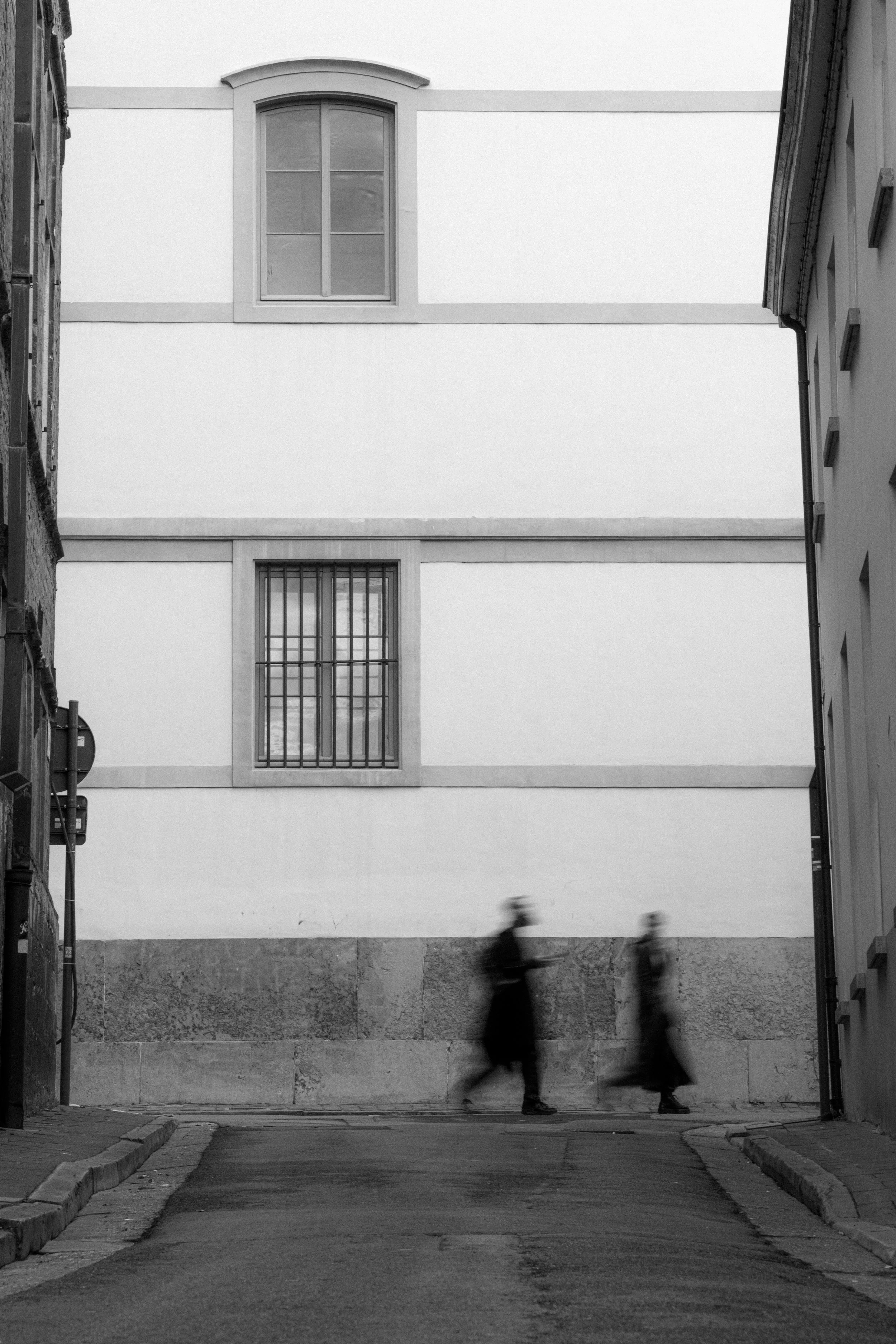 Black and white photo of two blurred pedestrians walking down a city sidewalk in front of a modern building with large windows and minimalist design.