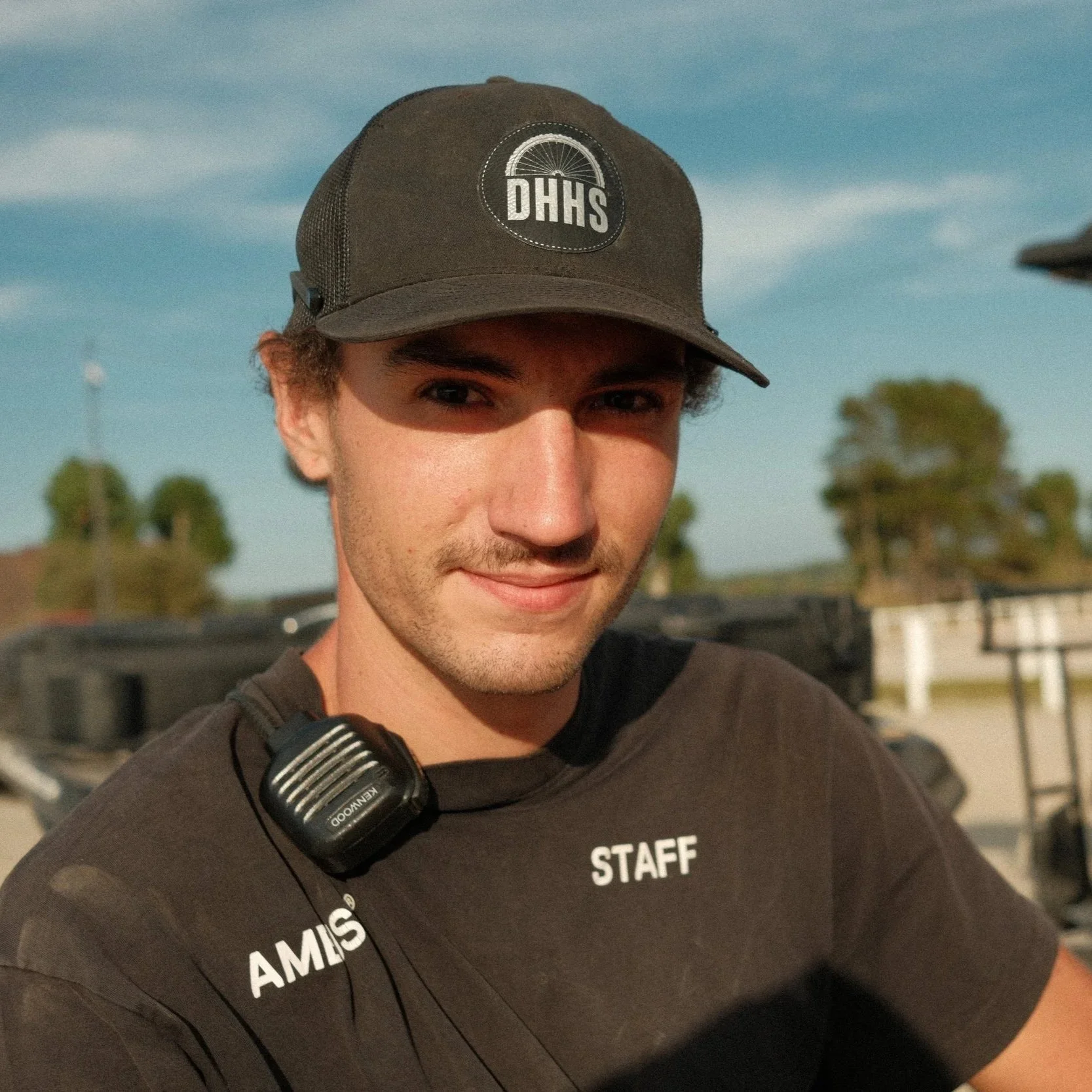 Close-up of a young man with brown hair, wearing a black 'STAFF' shirt and a black cap with a DHHS logo, standing outdoors under a blue sky with scattered clouds.