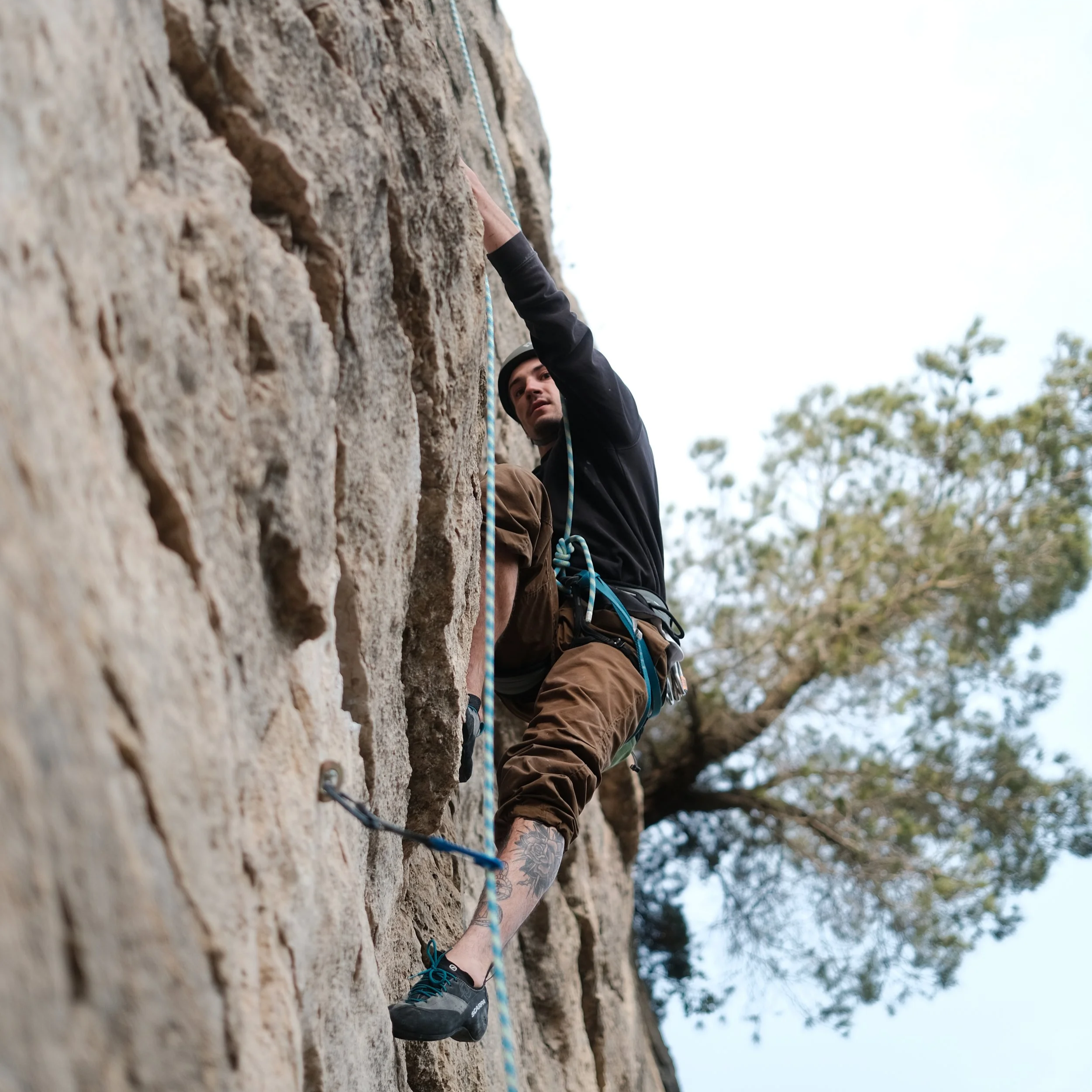 A person rock climbing on a vertical, textured stone wall outdoors, wearing climbing shoes, a black jacket, and a climbing helmet, with a blue safety rope.