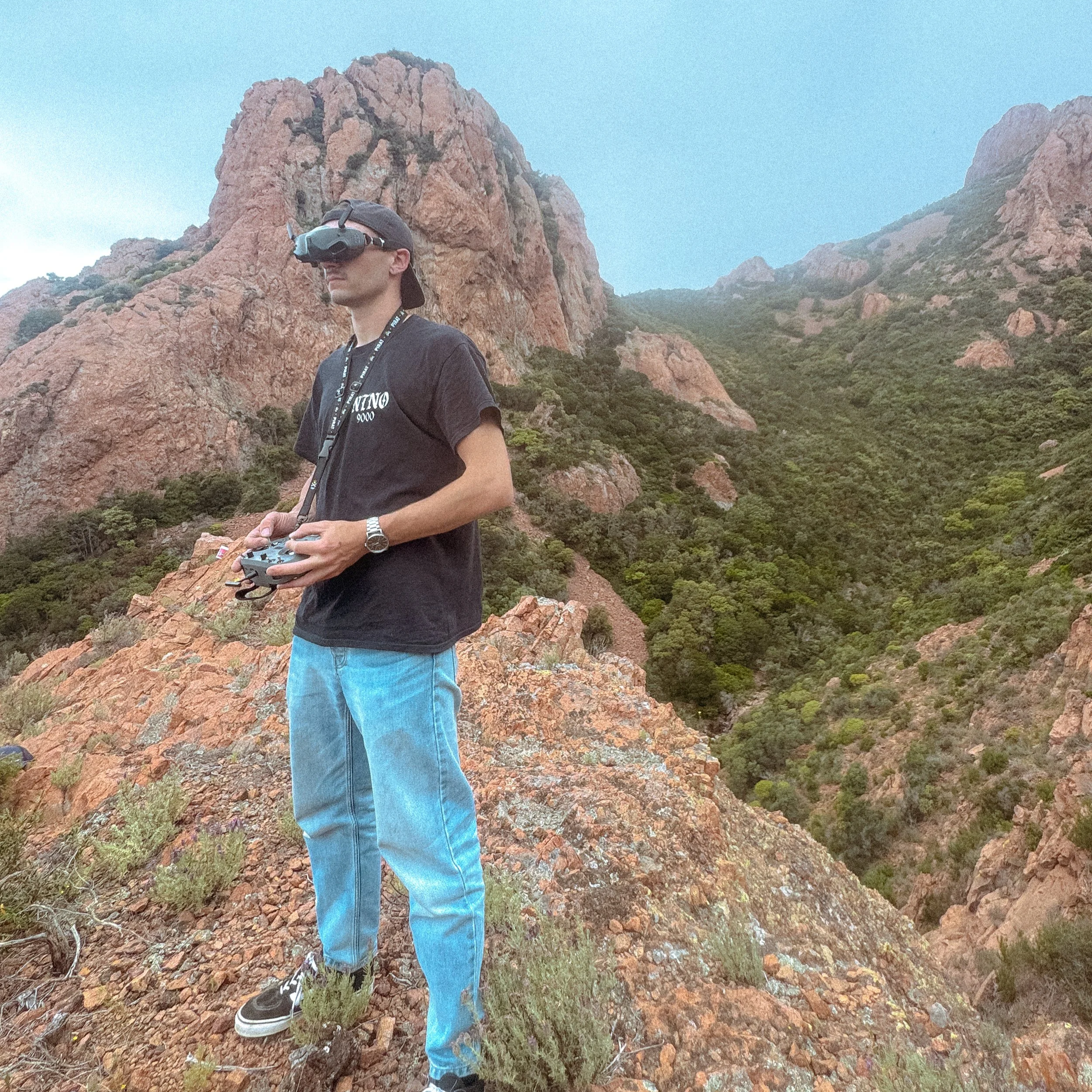 A young man with a black cap, sunglasses, black t-shirt, and blue jeans standing outdoors near rocky terrain and mountains, holding a remote control device, with a mountain peak in the background.