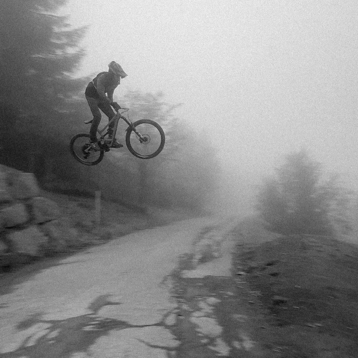 A person riding a mountain bike and performing a jump on a dirt trail in a foggy outdoor setting.