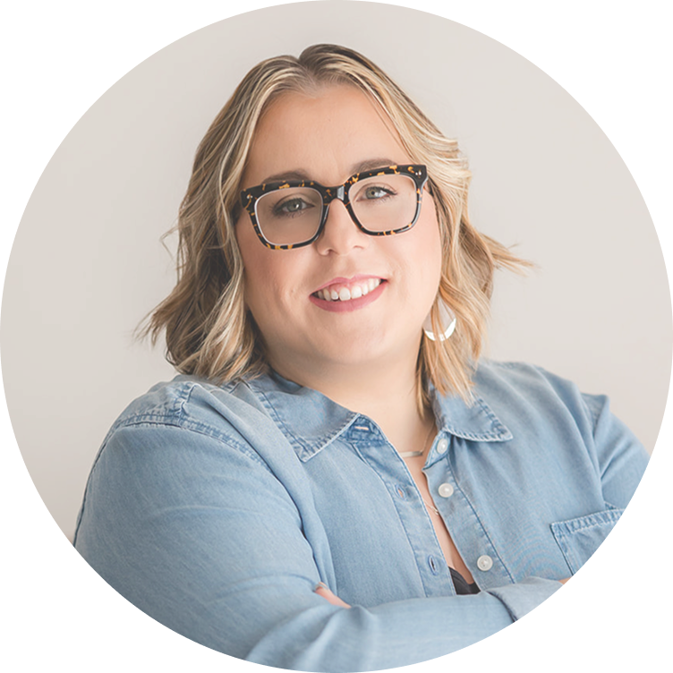 A portrait of Bridget Granholm who is smiling and has blonde hair wearing large tortoiseshell glasses and a light denim shirt