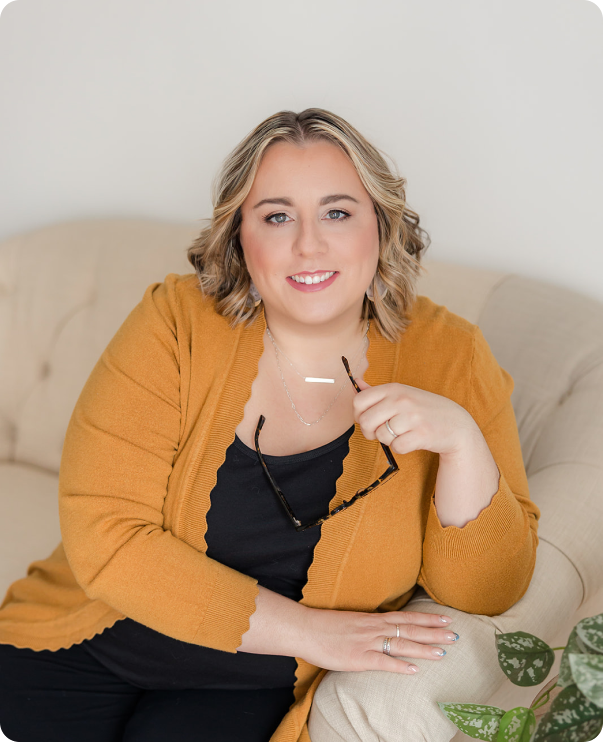 A portrait of Bridget Granholm smiling in her office: she has shoulder-length wavy blonde hair, wearing a mustard yellow cardigan over a black top, sitting on a beige couch, holding glasses.
