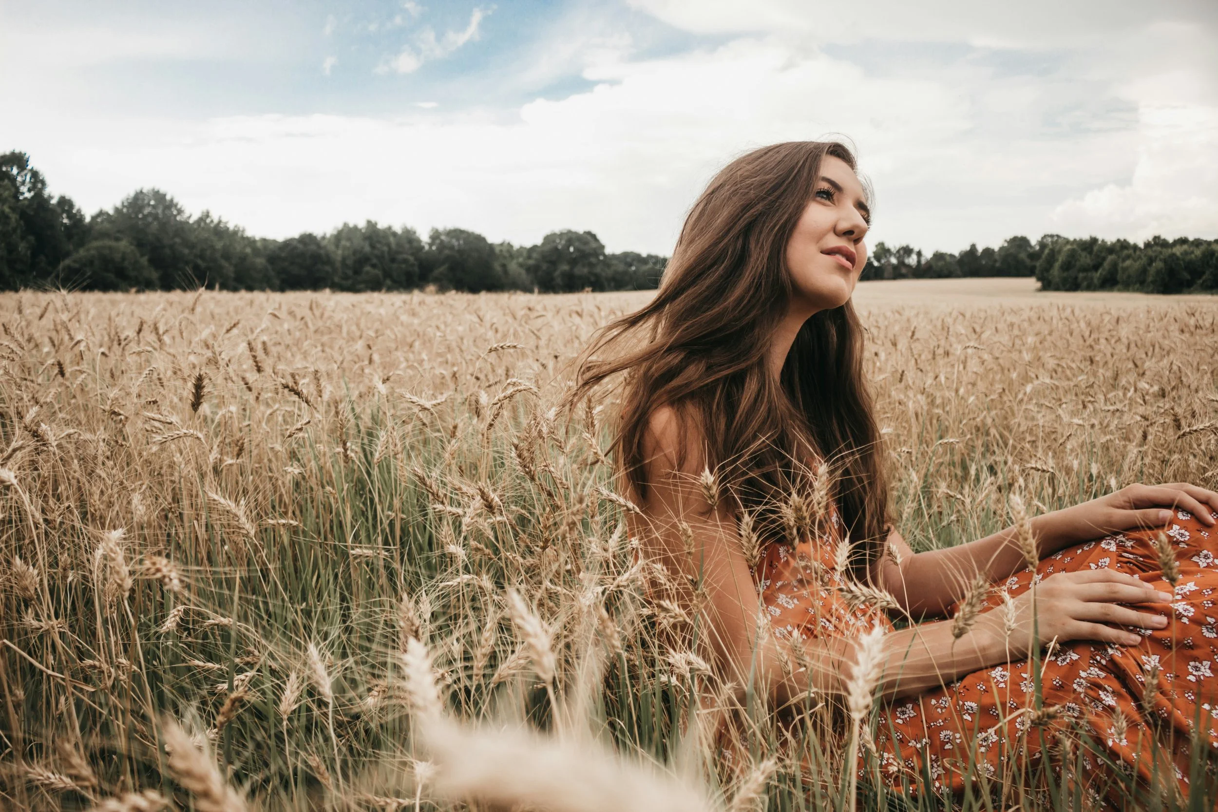 Young woman sitting in a wheat field with her eyes closed, wearing a floral dress and long brown hair, under a partly cloudy sky.