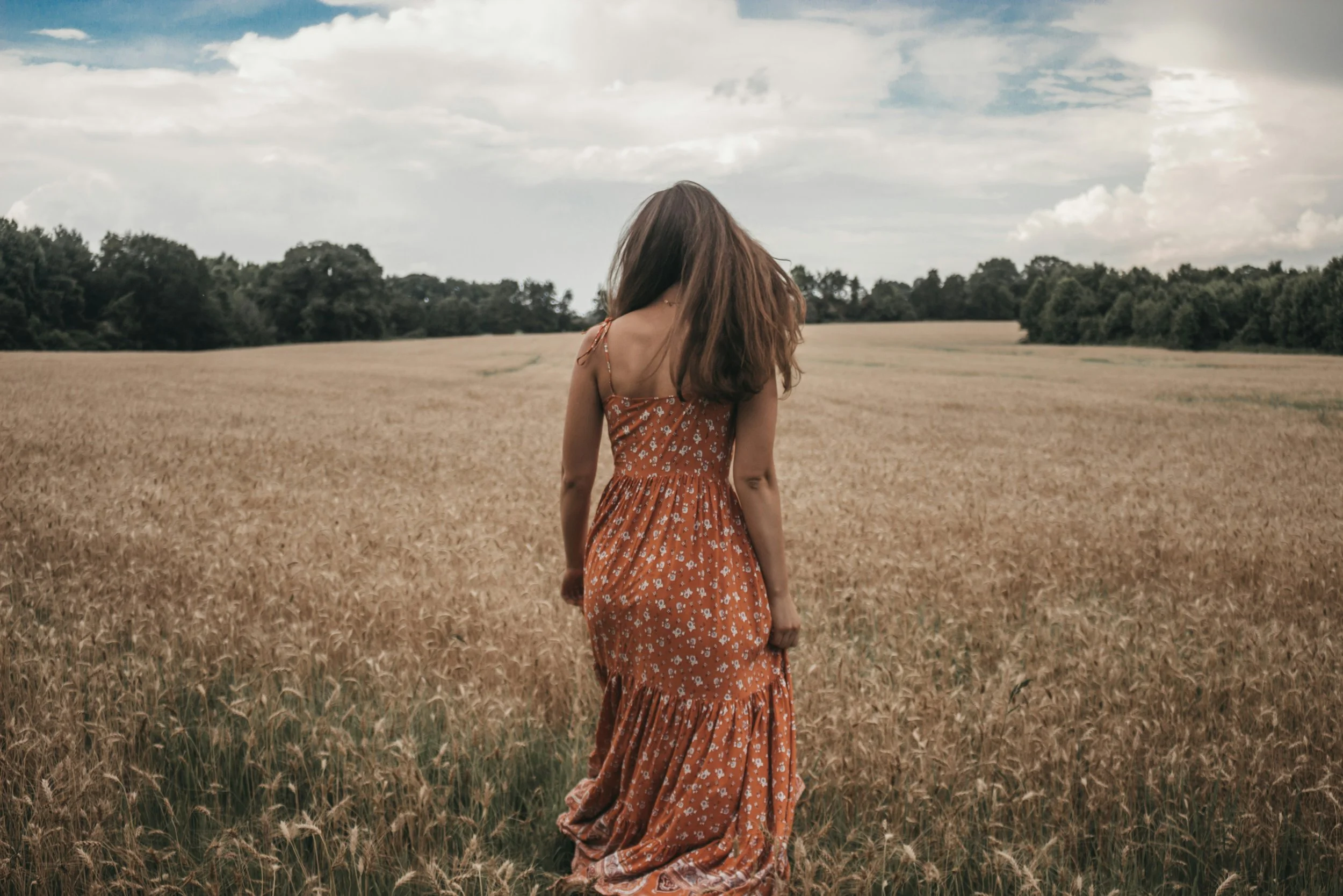 A woman in a long orange floral dress walking through a golden wheat field under a cloudy sky, with trees in the distance.