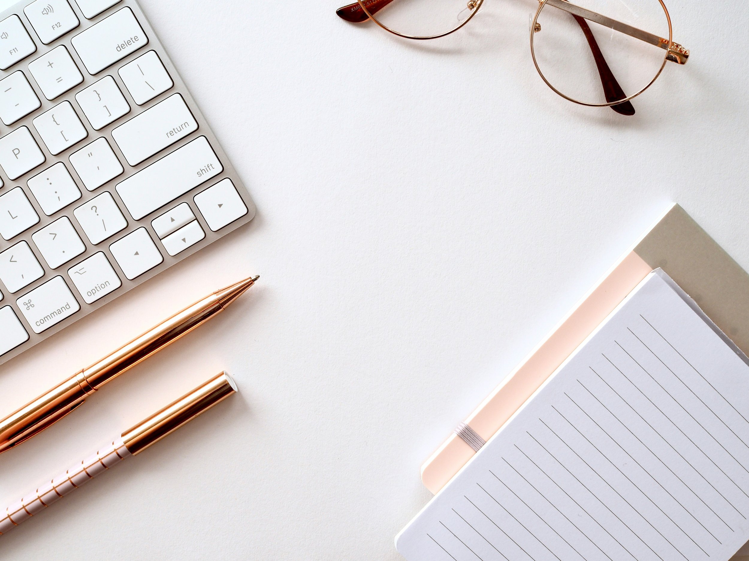 A white computer keyboard, a pair of rose gold sunglasses, a rose gold pen, a closed pink notebook, and a notebook with a grid pattern on a white surface.