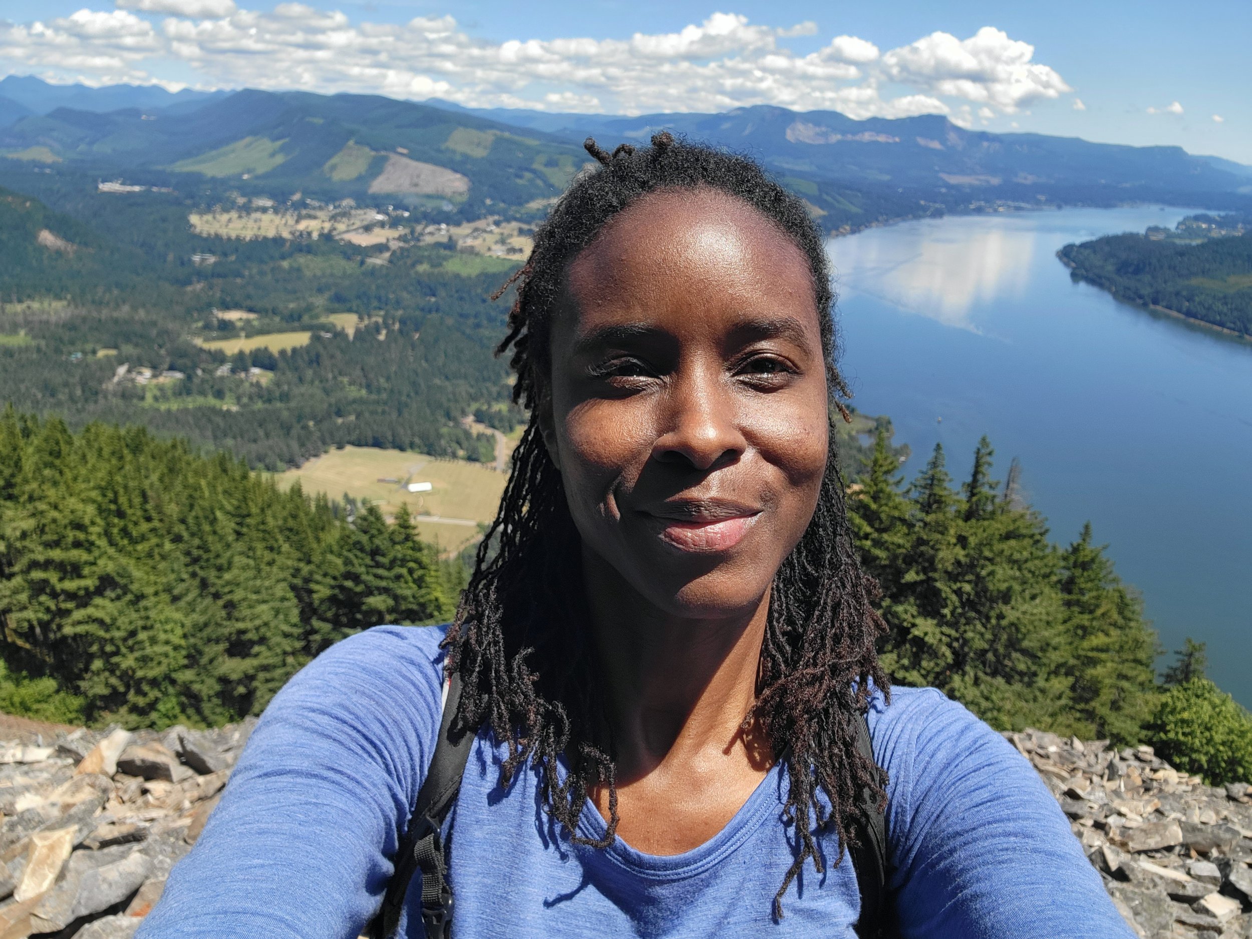 A woman smiling outdoors with a scenic view of mountains, a river, and a partly cloudy sky in the background.