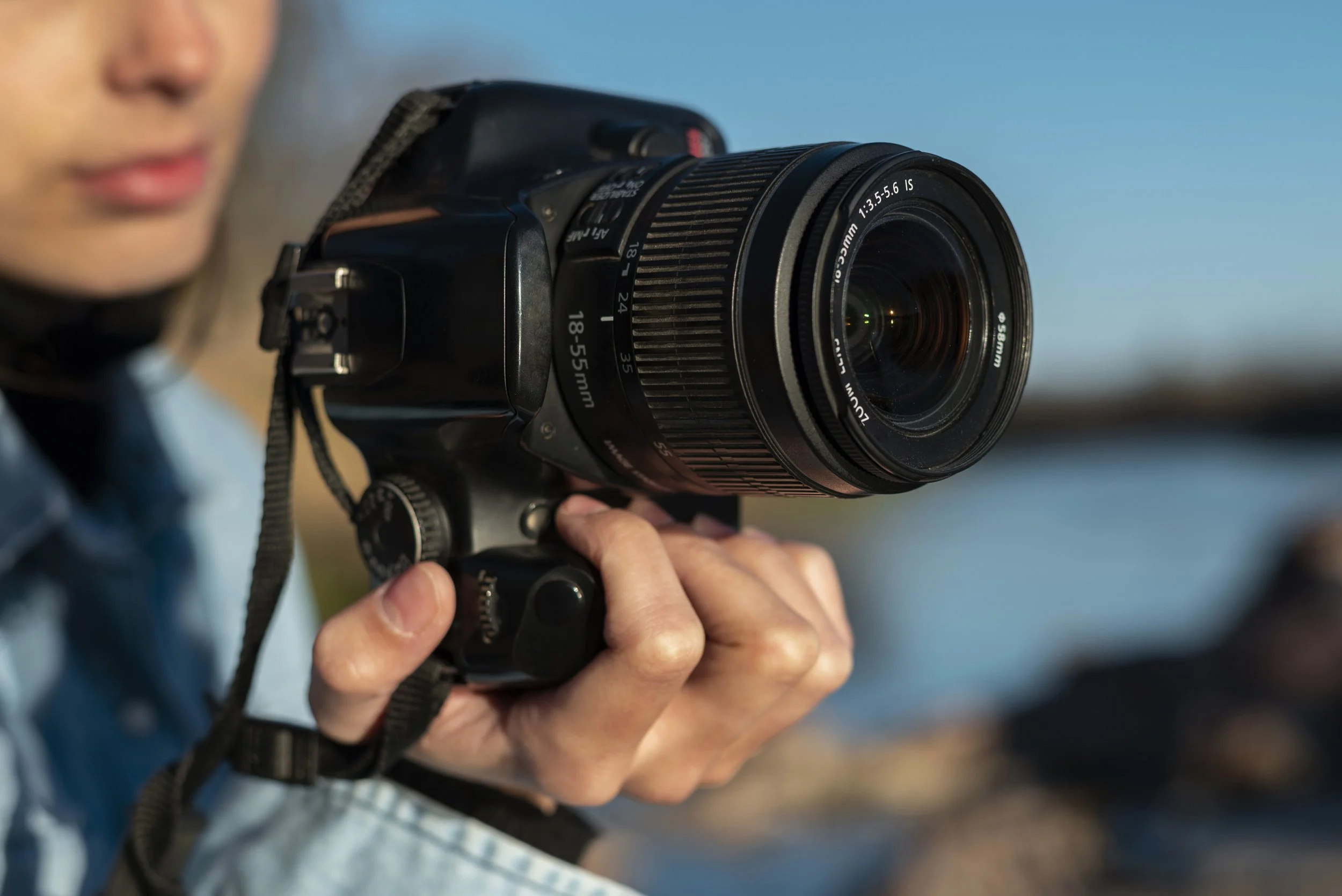 Personne tenant un appareil photo numérique avec un objectif de 18-55 mm, prise en extérieur avec un fond flou.