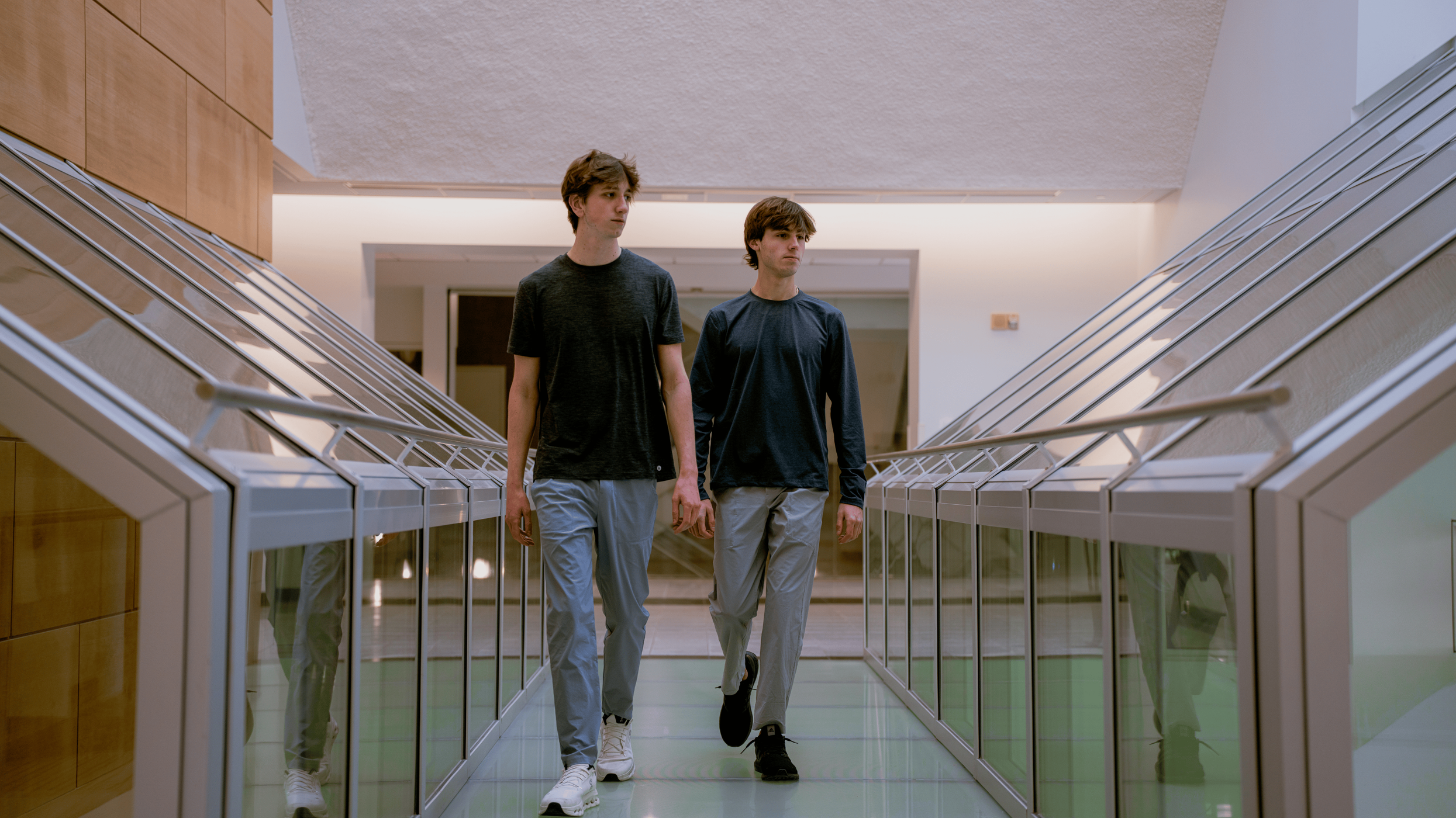 Two young men with brown hair walk side by side down a hallway with glass display cases and wood paneling on one side, and white walls with a textured ceiling.
