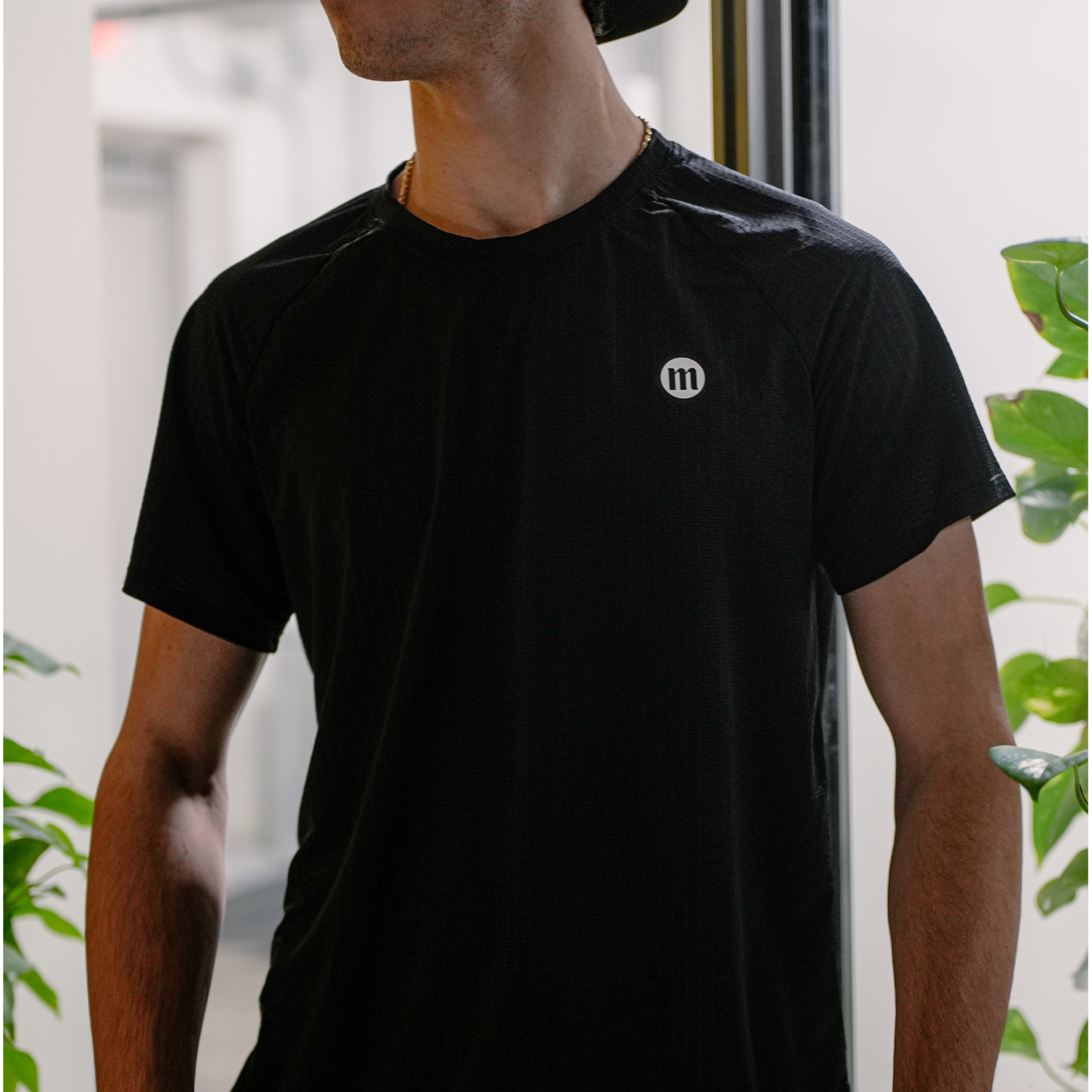 Close-up of a man wearing a black athletic shirt with a small white and black logo 'm' on the chest, standing indoors near green plants.