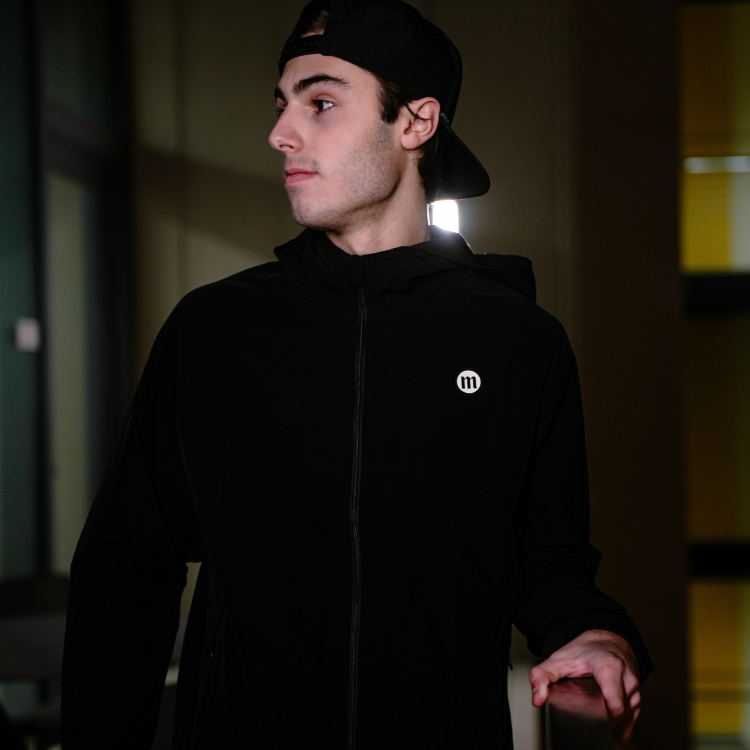 A young man wearing a black hoodie and backward cap, standing indoors with a side profile view, holding a suitcase handle.