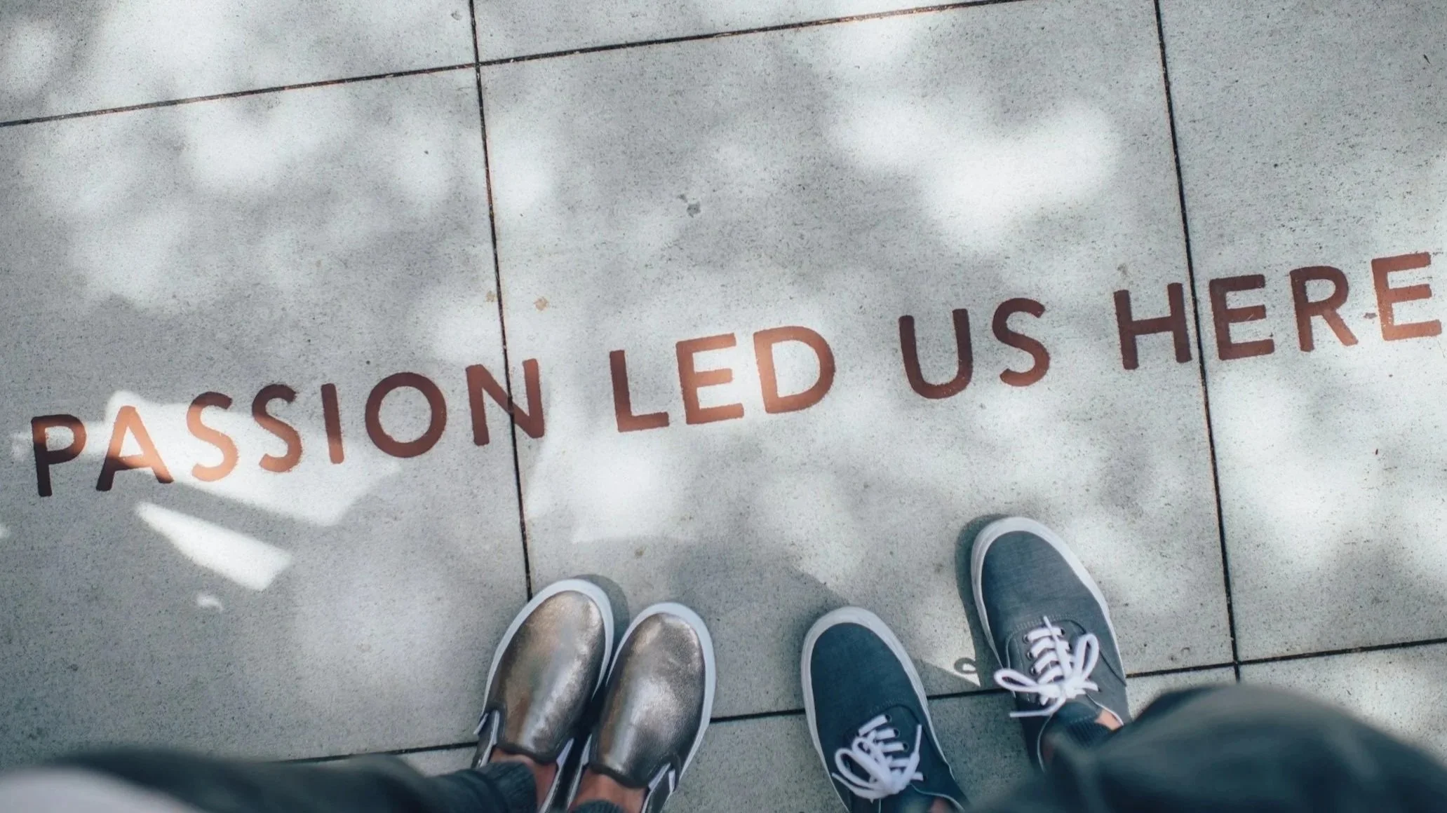 View from above of a tiled floor with the words "passion led us here" written on it. Two pairs of shoes are visible, one gold and one dark blue with white laces.