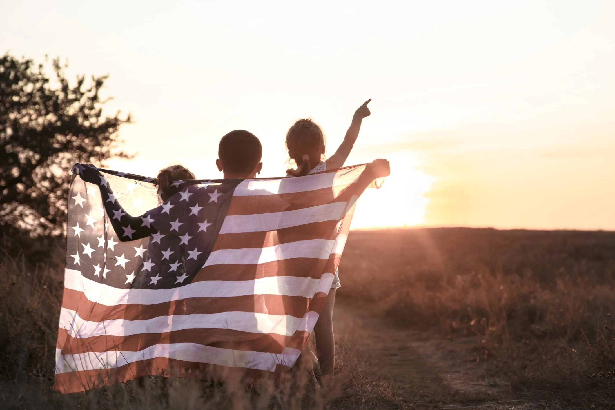 Three children walking on a dirt path at sunset, holding an American flag, with one child pointing towards the sky.