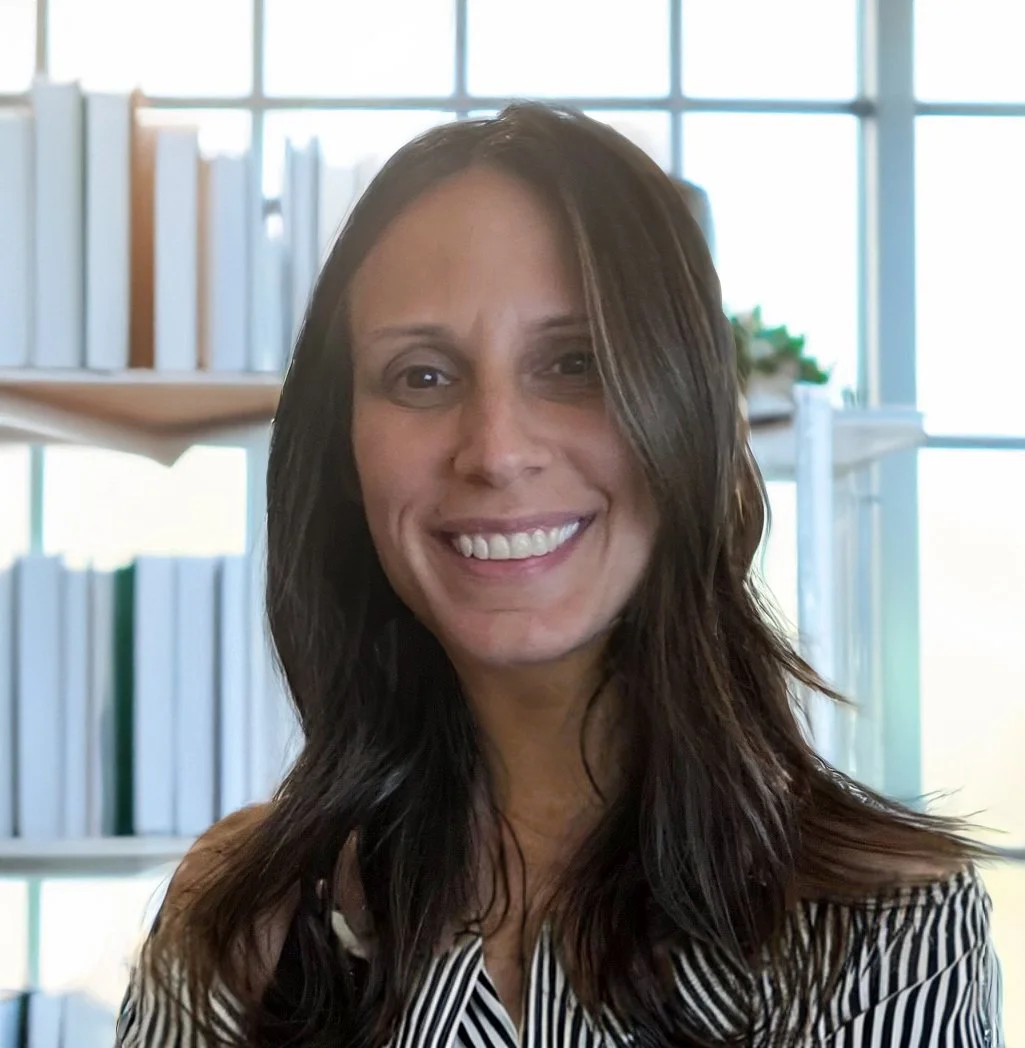 Smiling woman with long brown hair in an office with large windows and bookshelves in the background.