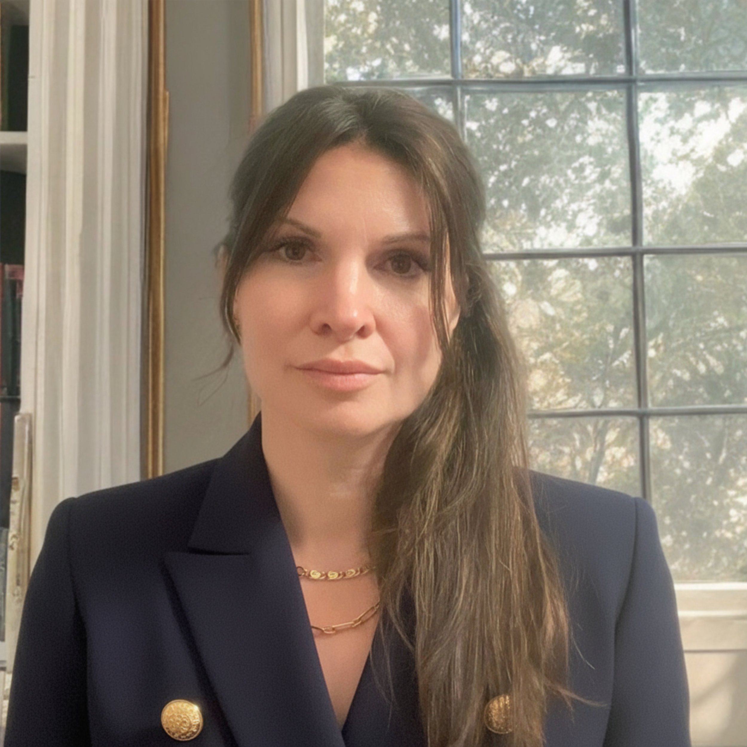 A woman with long brown hair wearing a dark blazer with gold buttons and gold necklaces, sitting in front of a window with trees outside.