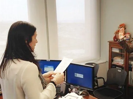 A woman with dark hair reading a paper in an office with two computer monitors on a desk.