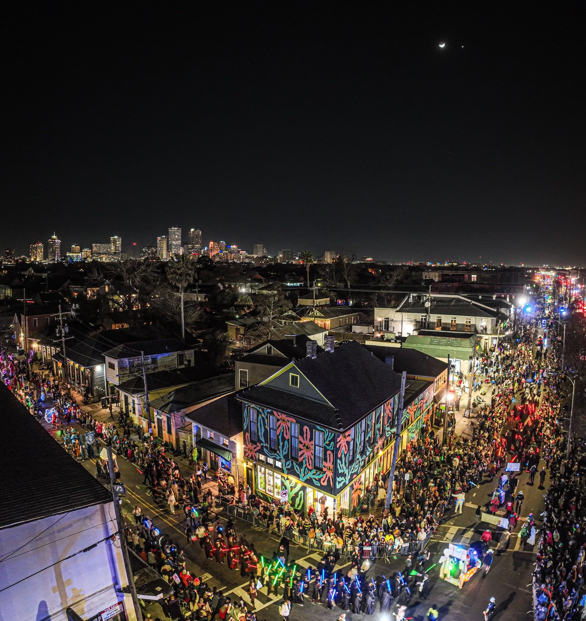 Nighttime cityscape with a crowd of people on the streets, a building with colorful mural art, and a skyline with tall buildings in the distance.