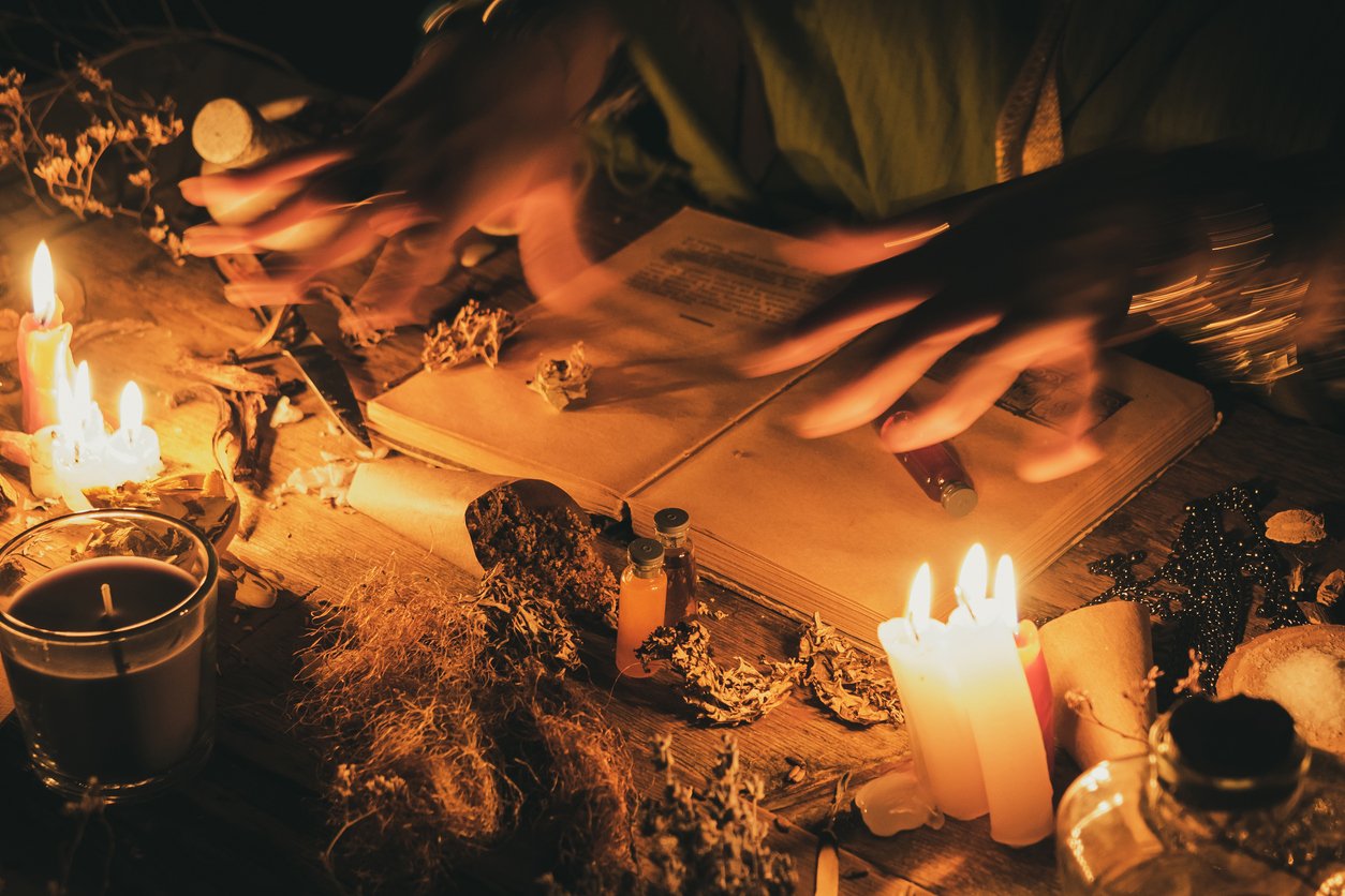 Person with blurred hands writing or reading in an open book at a dimly lit table with candles, herbs, jars, and spiritual objects.