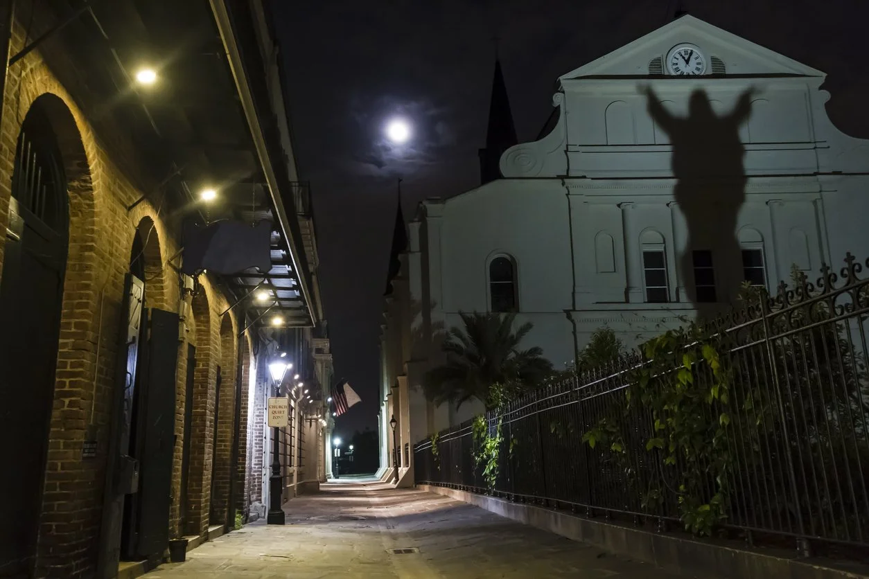 Nighttime scene on a city street with brick buildings on the left, a church with a clock on the right, a moon shining in the cloudy sky, a shadow of a person with arms raised cast on the church, and a fence with plants along the sidewalk.