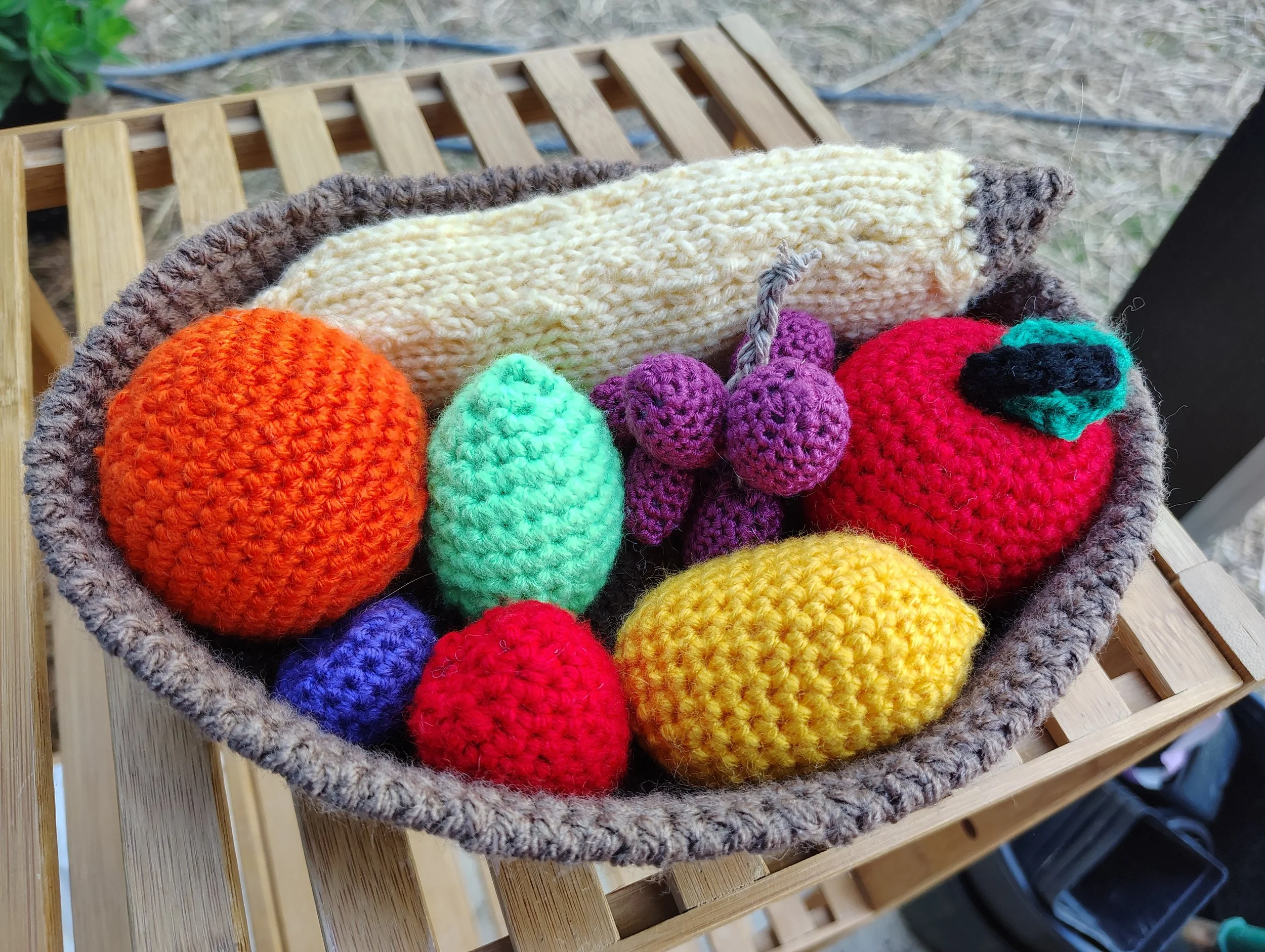 brown crochet basket with crochet and knit fruits. There's an orange, an apple, a banana, a lime, a lemon, a small bunch of grapes, a plum, and a strawberry.