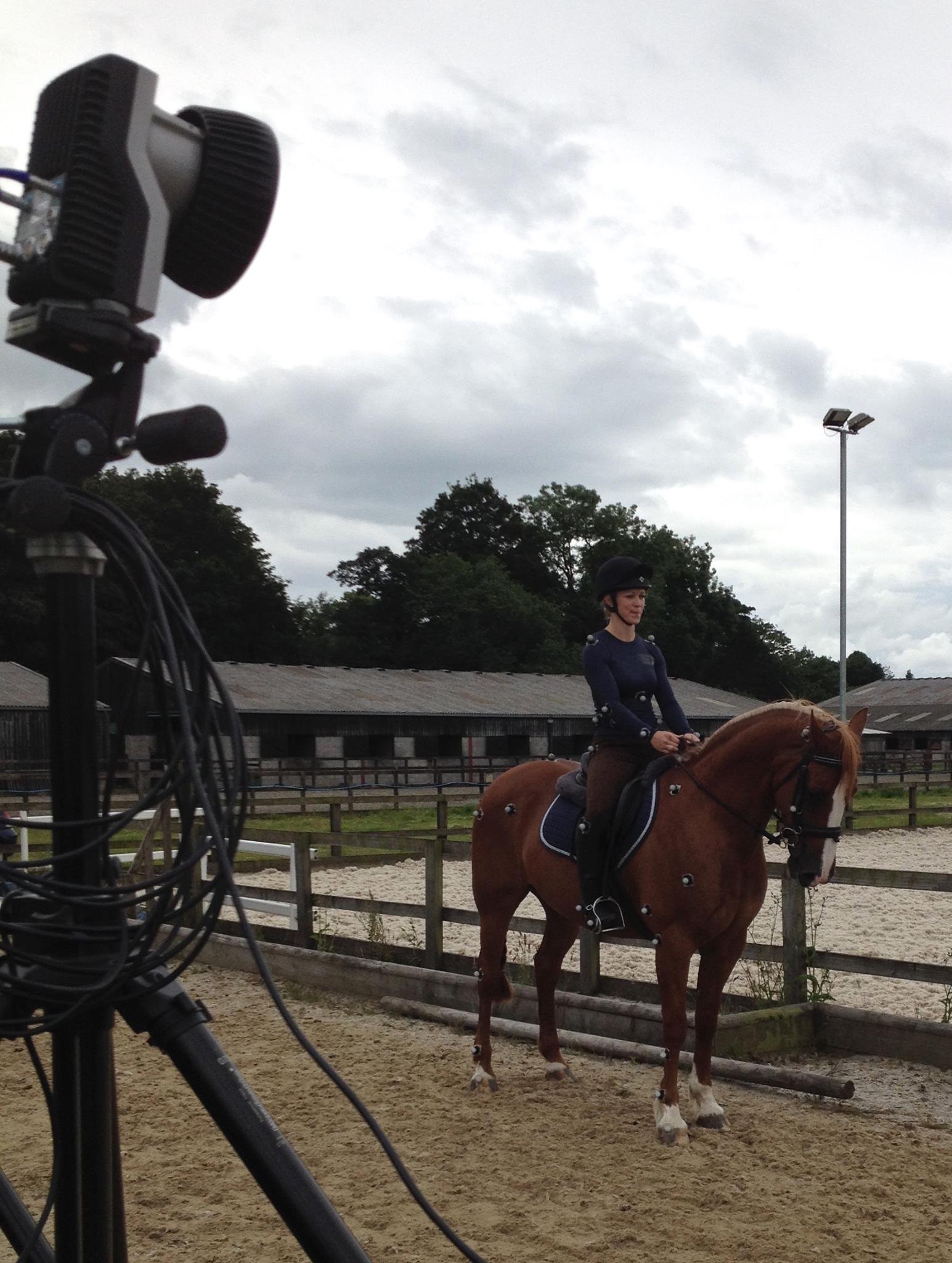 A woman riding a brown horse in an outdoor riding arena, with a large optical motion capture (qualisys) camera on a tripod in the foreground and cloudy sky above. The woman and horse have retroreflective markers attached over anatomical landmarks.