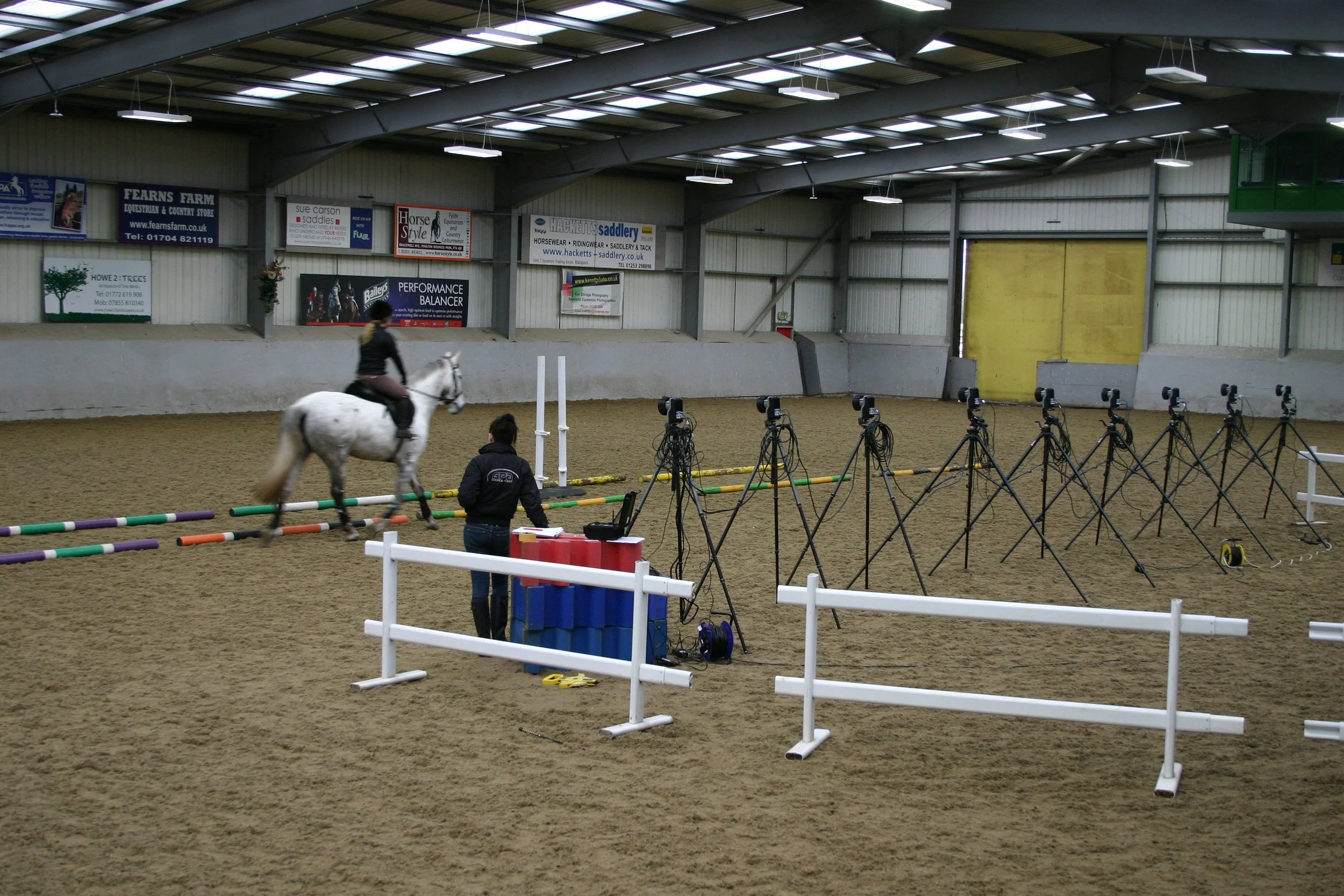 Indoor equestrian arena with a person riding a white horse in front of a qualisys optical motion capture system. Another person operates the laptop and camera system.
