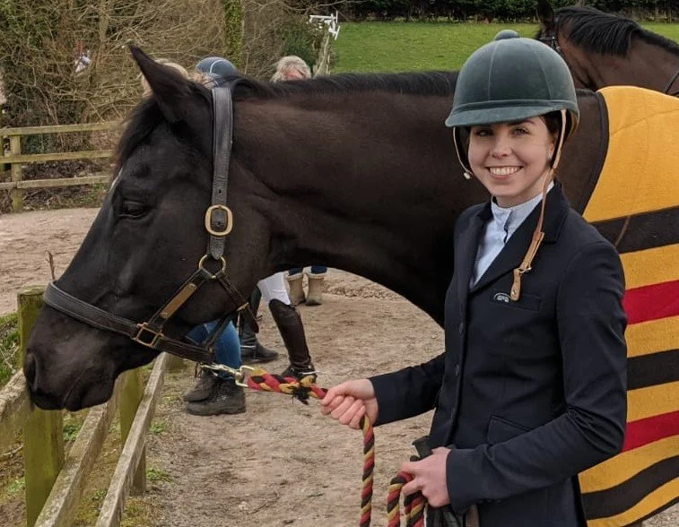 A young woman in riding gear holding a black horse's reins at a competition, with other people and horses visible in the background.