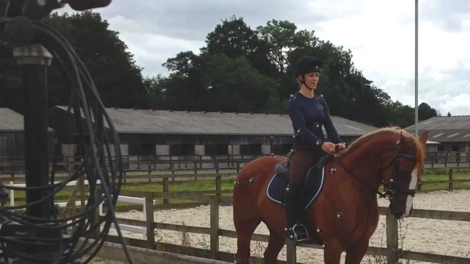 A woman riding a horse in an outdoor riding arena, with stable buildings and trees in the background, under a cloudy sky.