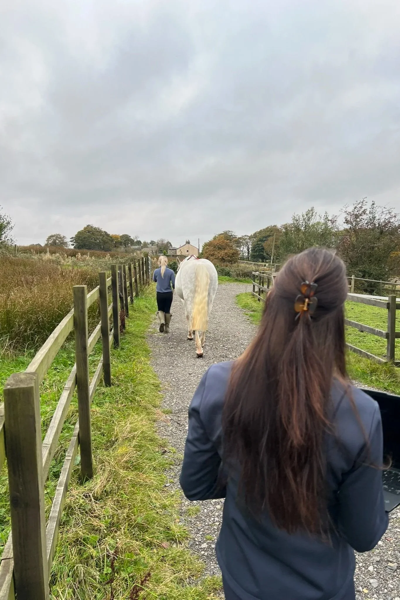 A person with long hair in a ponytail and a navy jacket is walking on a gravel path beside a wooden fence, with a woman leading a horse ahead. The scene appears to be in a rural area with grassy fields, trees, a cloudy sky, and distant houses.