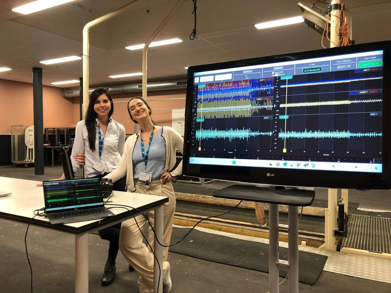 Two women standing behind a table with a Delsys Trigno surface electromyography system, smiling at the camera, in front of large monitors displaying equine sEMG waveforms and data, in a clinical setting with an equine treadmill. 