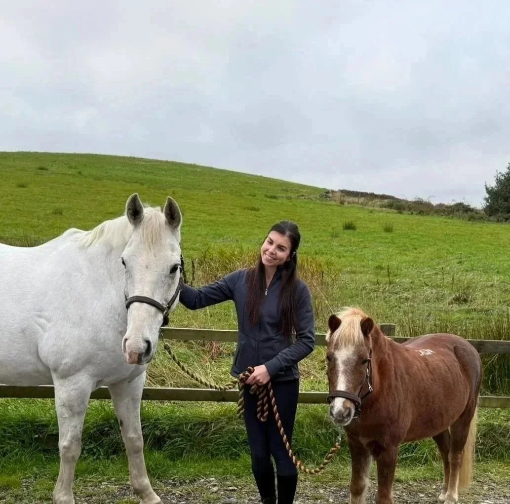 A young woman with long dark hair smiling and holding two small horses, one white and one brown, on a grassy field with a hill and cloudy sky in the background.