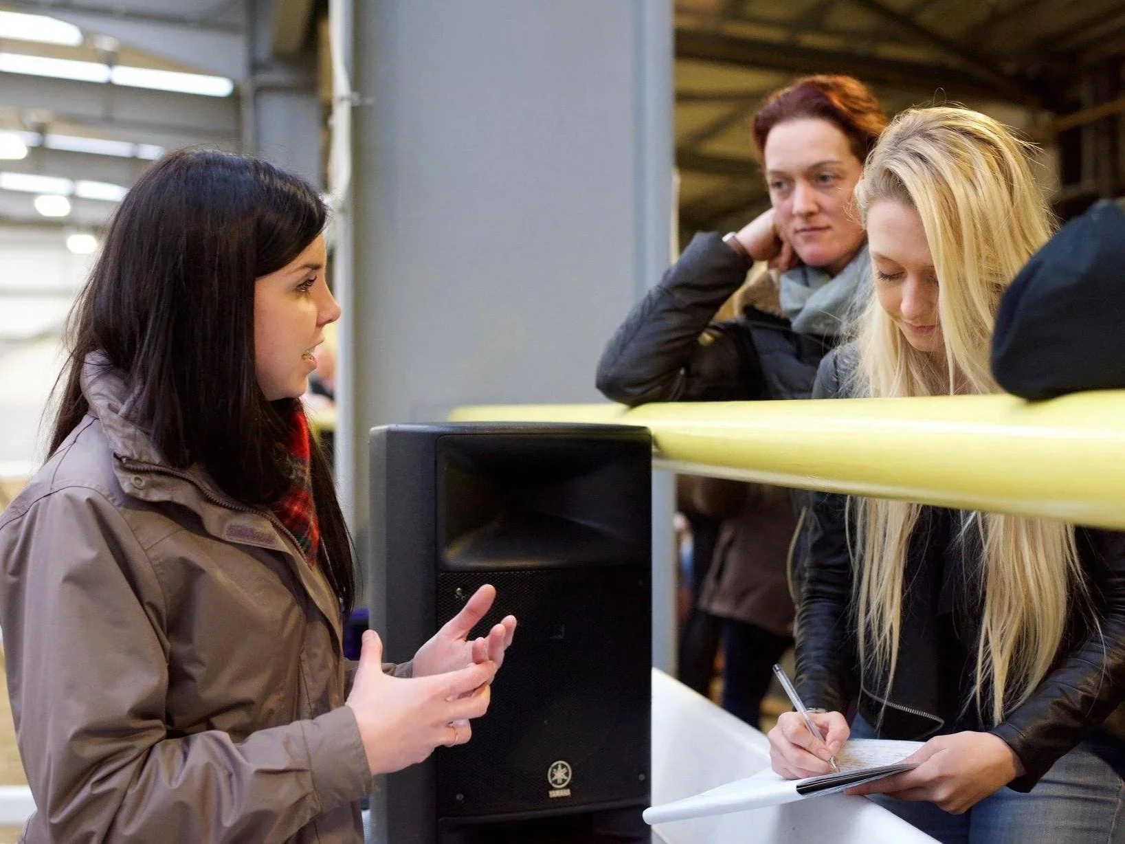 A woman answers equine biomechanics questions to two members of the audience at an educational workshop. One of the listeners is taking notes.