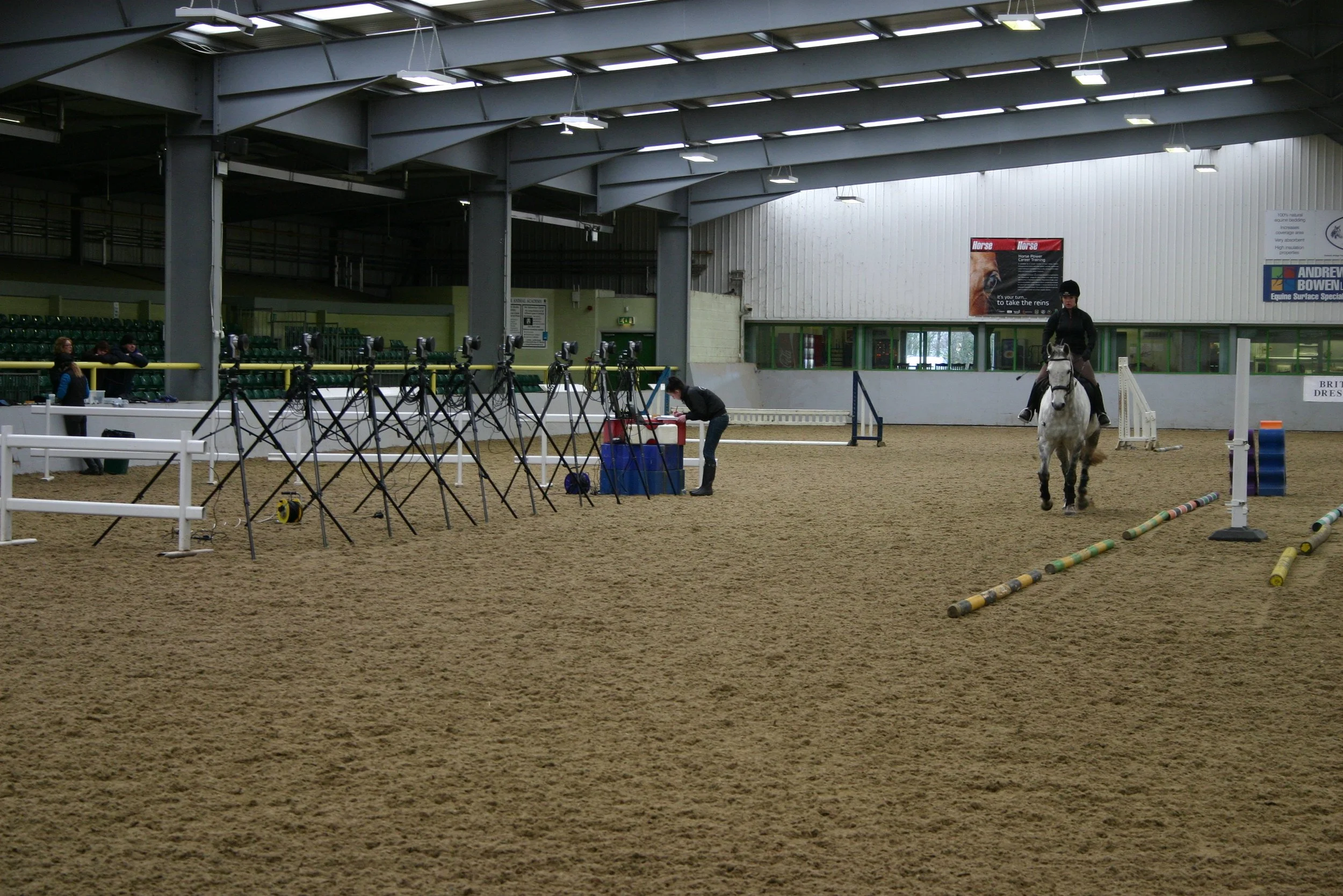An indoor equestrian arena with an optical motion capture system (qualisys) set up with cameras on tripods. There is a person on horseback and a person at a laptop collecting 3D equine biomehanics / kinematic data from the horse trotting.