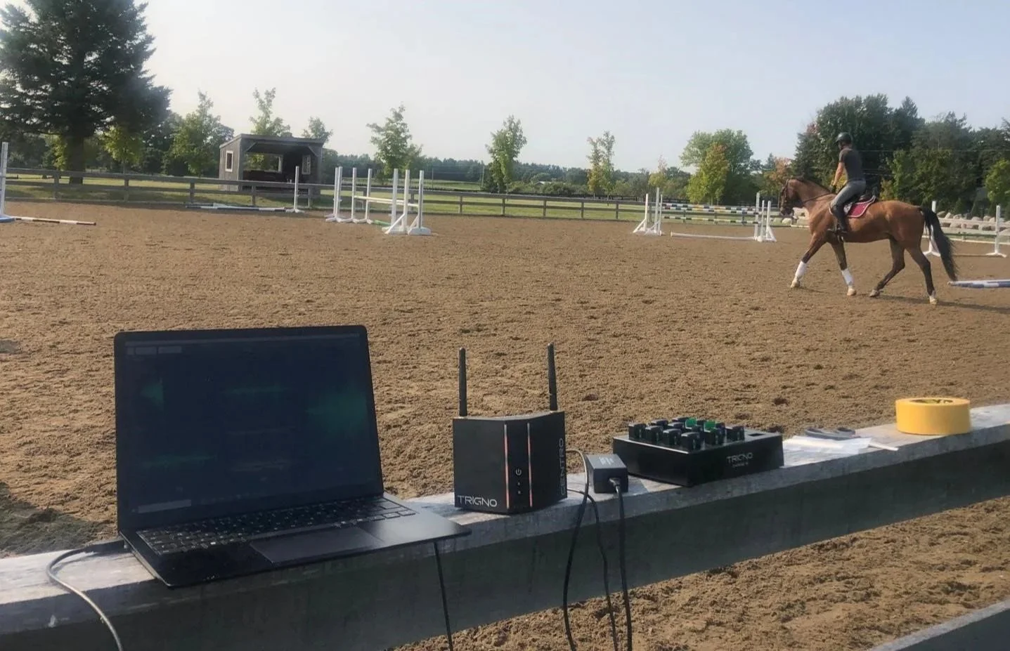 An outdoor equestrian jumping arena with a rider on a brown horse practicing. Equipment such as a laptop and Delsys surface electromyography system are set up on a ledge in the foreground.